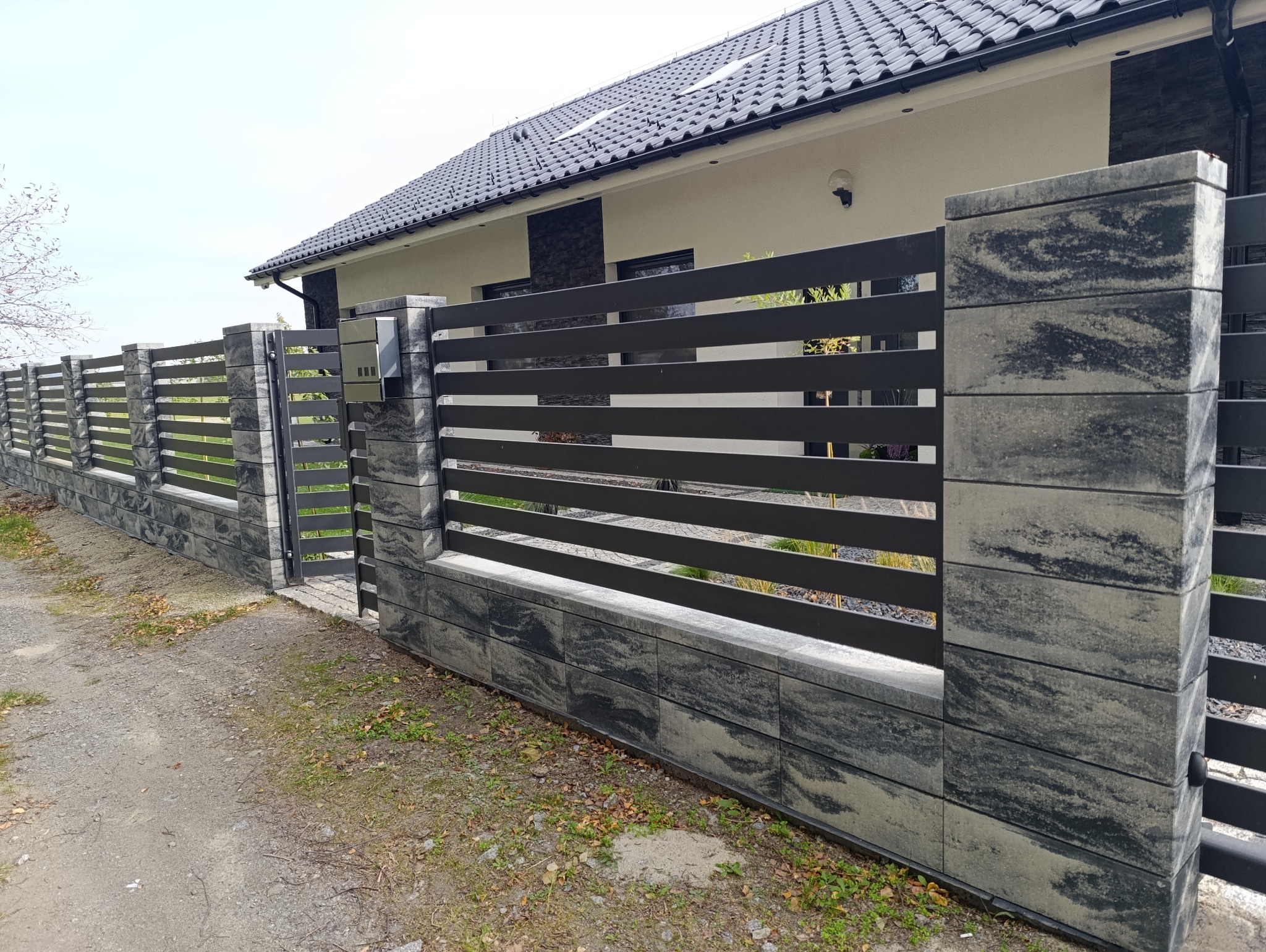 Nowoczesne ogrodzenie z betonowych bloczków o marmurkowym wzorze i poziomych, czarnych przęseł. Widoczna furtka i skrzynka pocztowa, dom w tle.