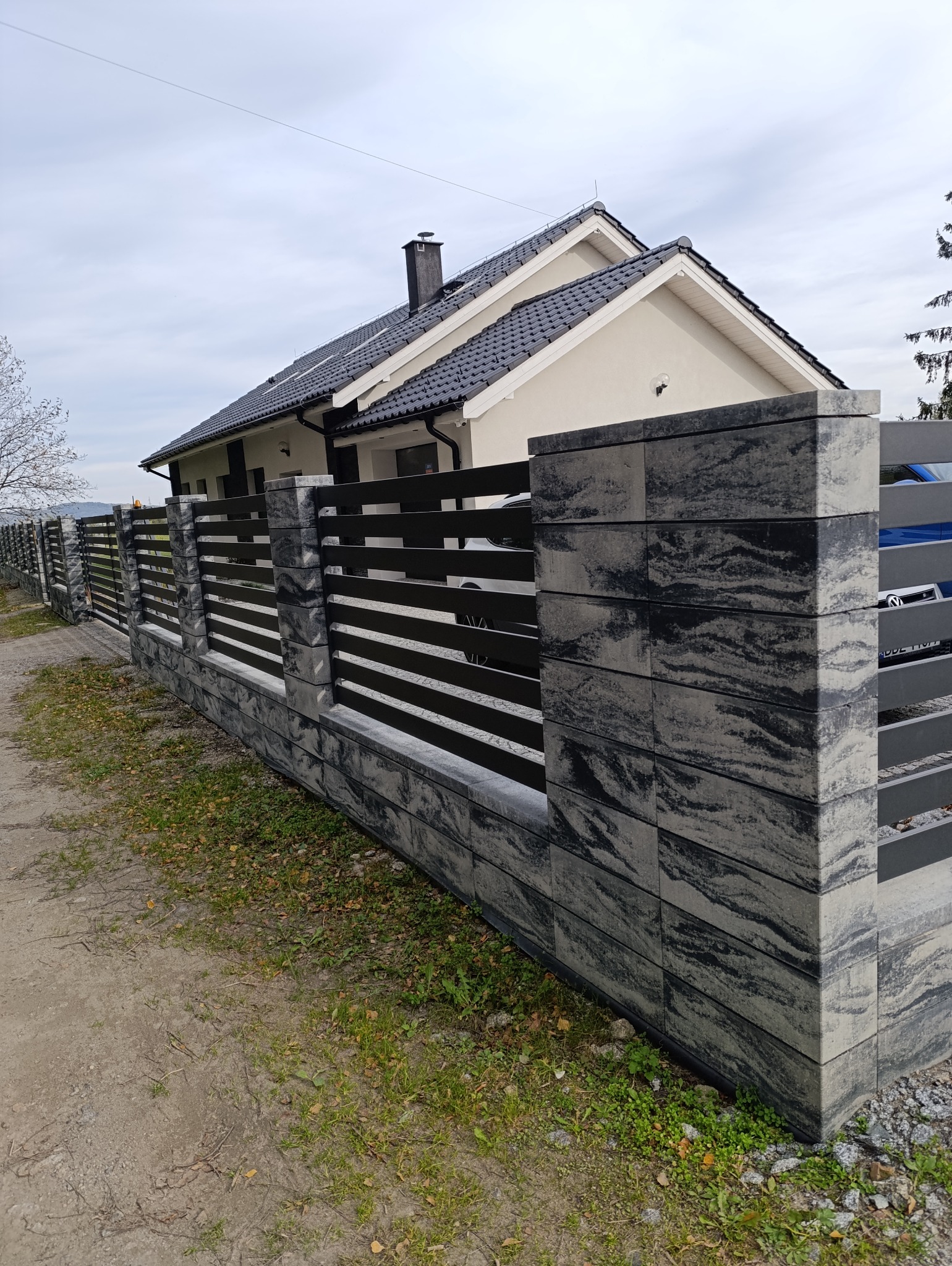 Nowoczesne ogrodzenie z betonowych bloczków w odcieniach szarości i czarnych, poziomych przęseł, na tle domu z ciemną dachówką w pochmurny dzień.