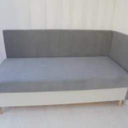 Szara tapicerowana kanapa narożna z pionowymi przeszyciami na oparciu, umieszczona na białej podstawie z widocznymi drewnianymi nóżkami.