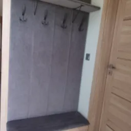 Tapicerowany panel ścienny z wieszakami i siedziskiem w przedpokoju, jasne drewno, szara tkanina, minimalistyczny design, obok drewniane drzwi.