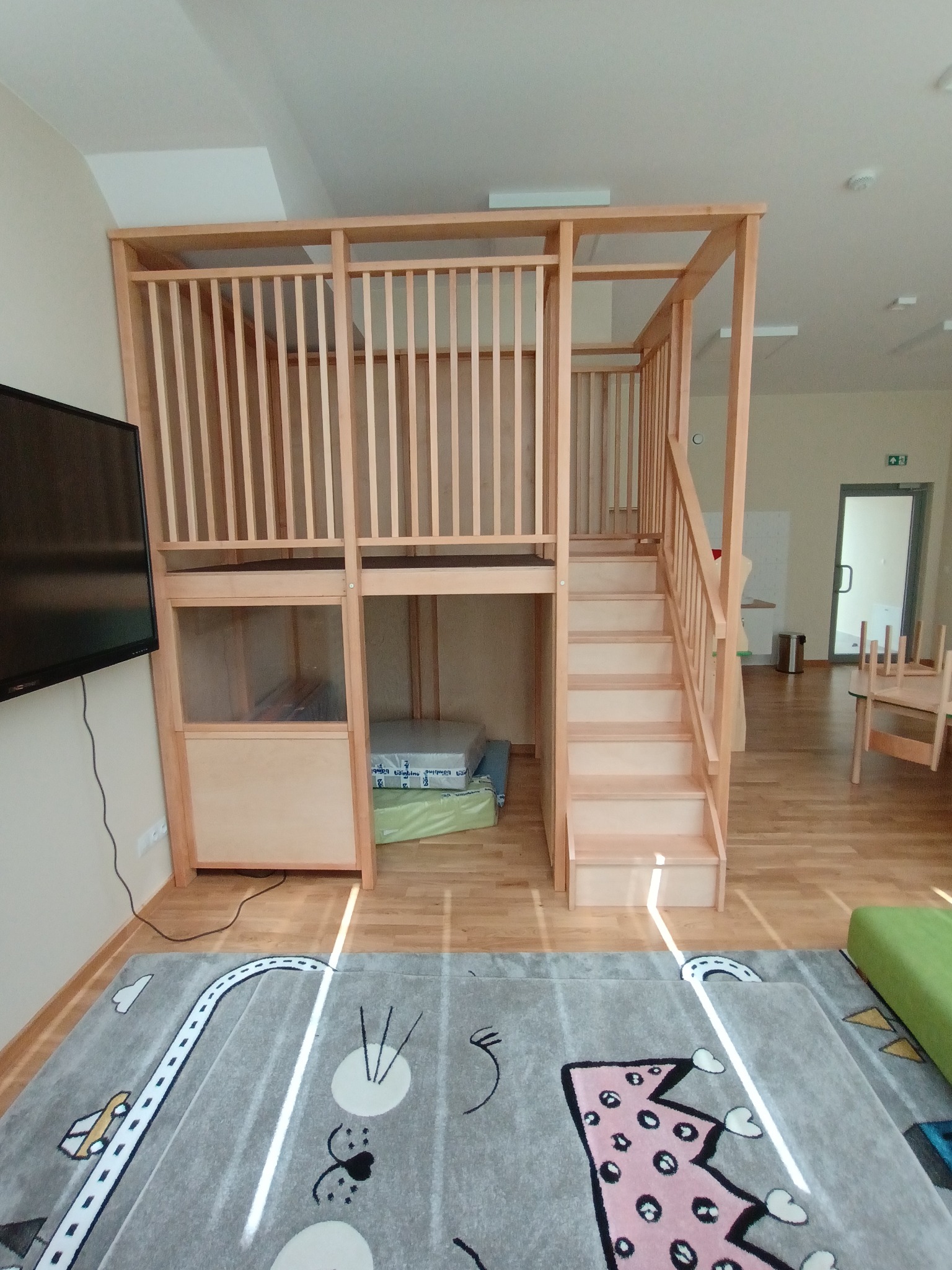 Drewniana antresola dla dziecka z drabinką, umieszczona w pokoju zabaw z dywanem w motywy drogowe.