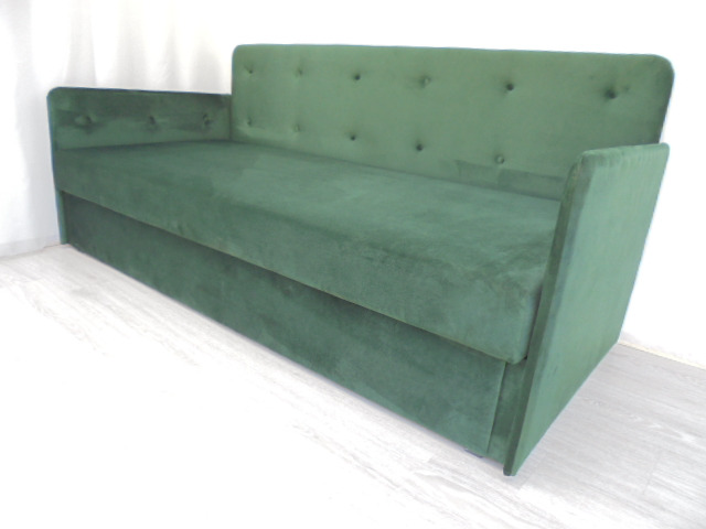 Rozkładana sofa w odcieniach butelkowej zieleni, obita miękkim welurem, z ozdobnymi guzikami na oparciu i bokach, ustawiona na jasnej podłodze.