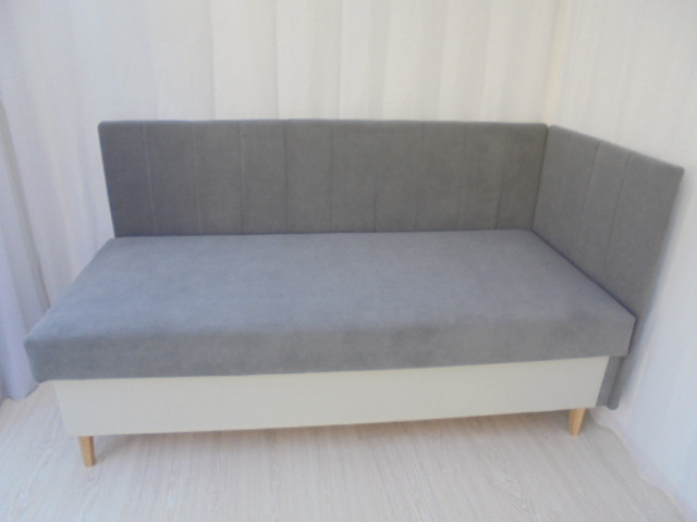 Szara tapicerowana kanapa narożna z pionowymi przeszyciami na oparciu, umieszczona na białej podstawie z widocznymi drewnianymi nóżkami.