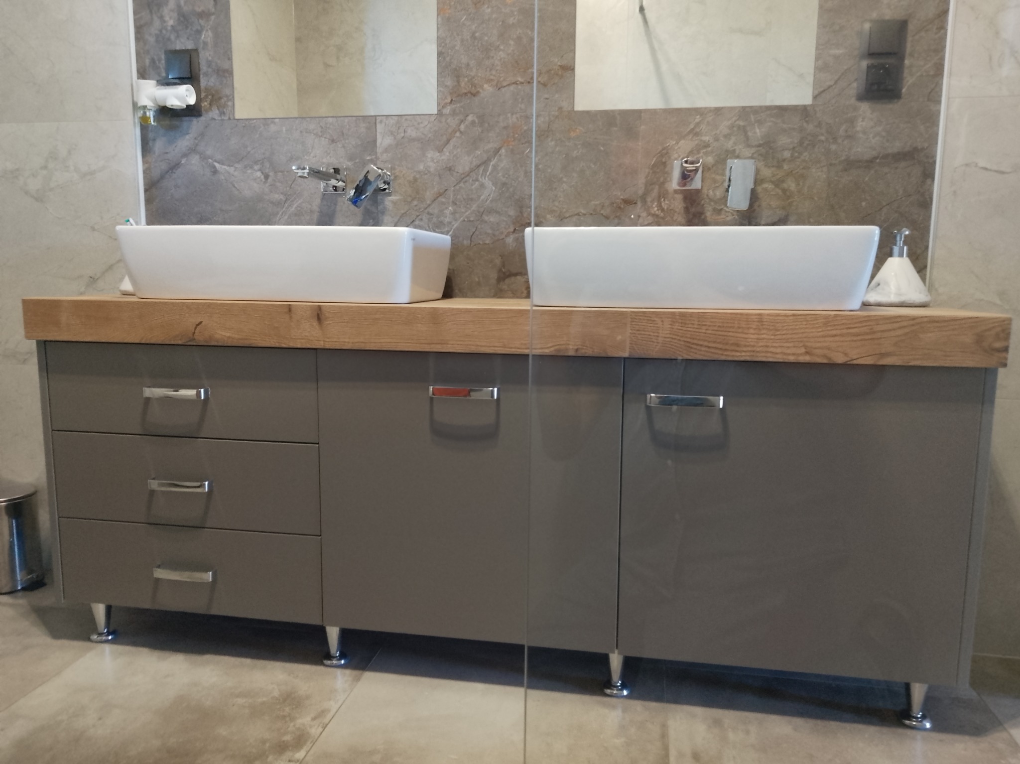 Szara szafka łazienkowa z drewnianym blatem i dwiema białymi umywalkami nablatowymi, chromowane nóżki i uchwyty. Minimalistyczny design, marmurowe płytki w tle.