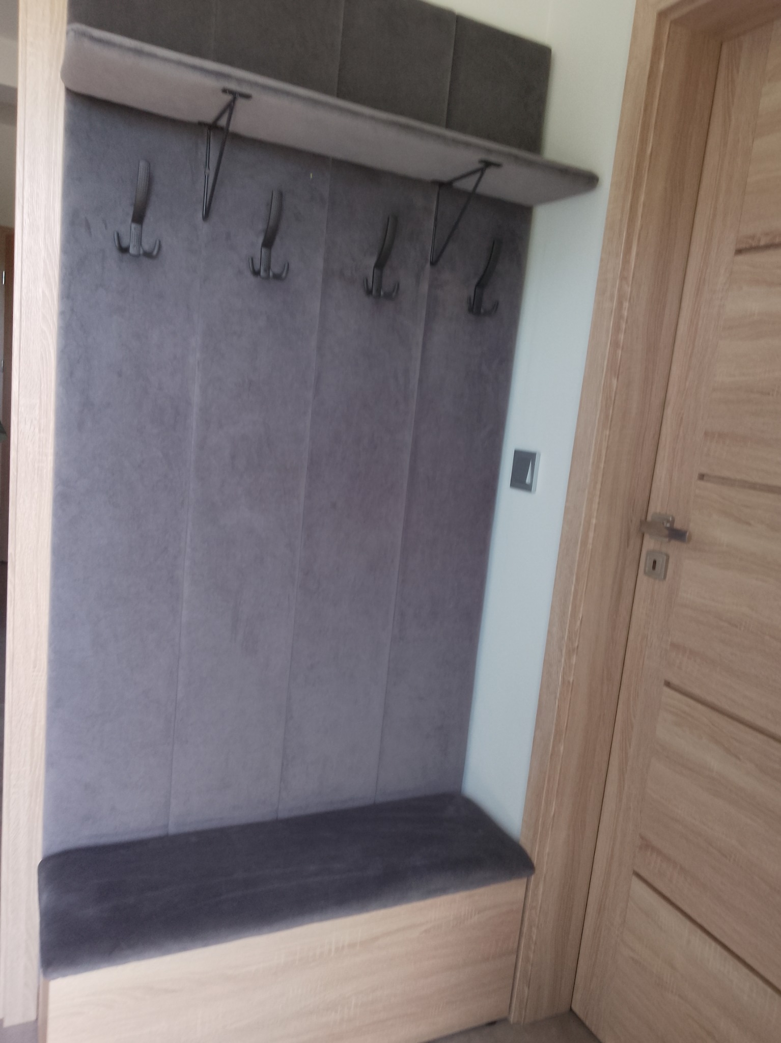 Tapicerowany panel ścienny z wieszakami i siedziskiem w przedpokoju, jasne drewno, szara tkanina, minimalistyczny design, obok drewniane drzwi.
