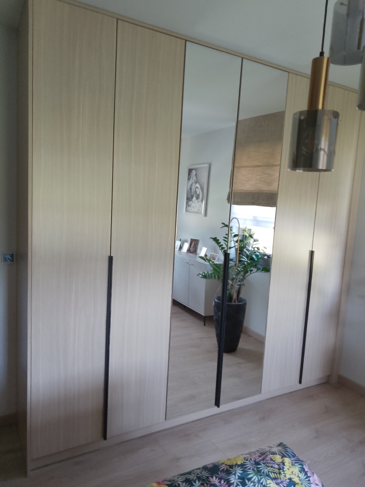 Jasna szafa z lustrami wkomponowana w przestrzeń pokoju, odbijająca wnętrze z rośliną doniczkową i komodą. Proste, czarne uchwyty.
