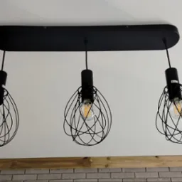 Czarna, designerska lampa sufitowa z trzema żarówkami w metalowych kloszach o nieregularnym kształcie, zamontowana na białym suficie. U dołu widoczny fragment ceglanej ściany.