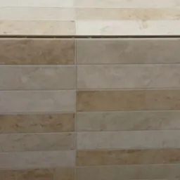 Ściana wyłożona beżowo-brązowymi, prostokątnymi płytkami ceramicznymi, z użyciem klipsów poziomujących. Widoczne fugi i fragment pomarańczowej taśmy malarskiej u góry.