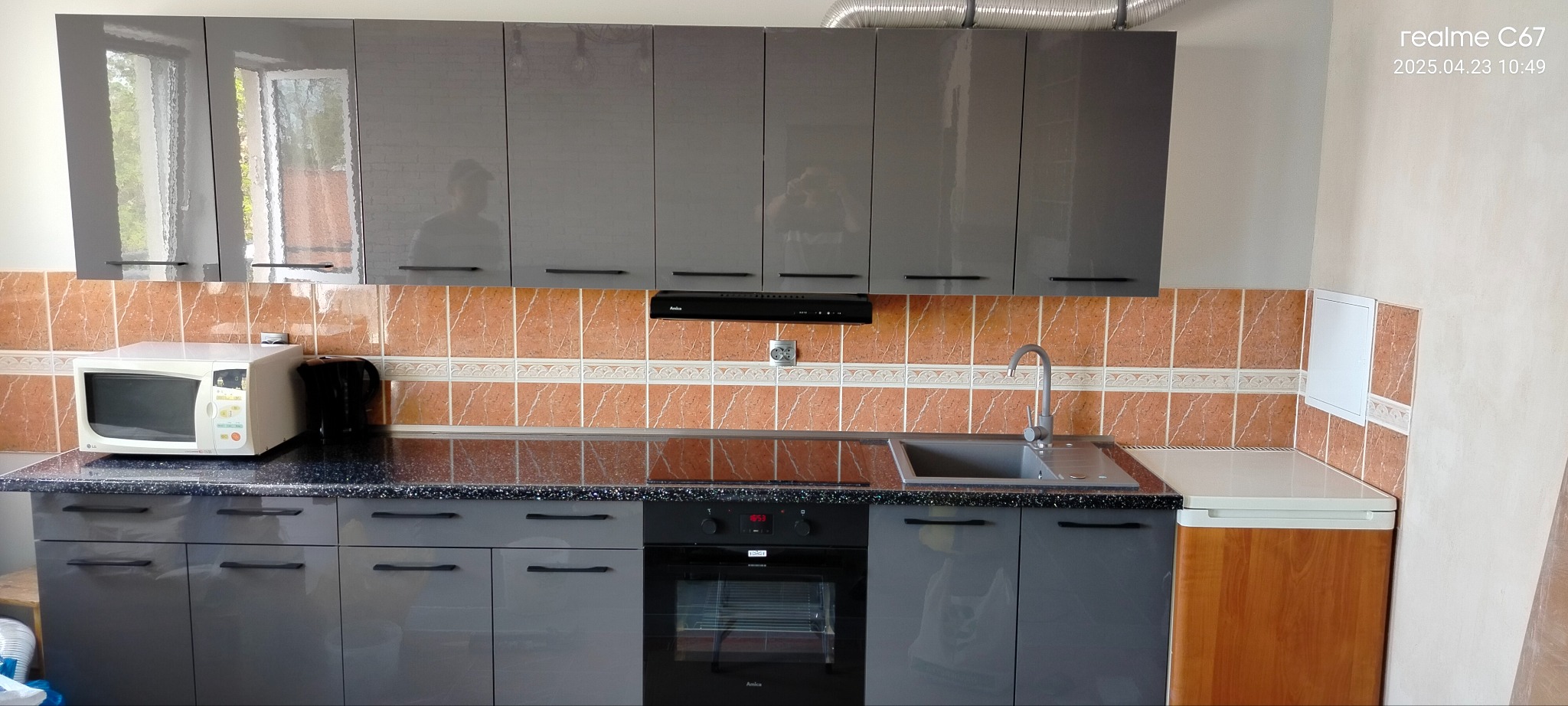 Nowoczesna kuchnia: szare szafki z czarnymi uchwytami, brązowe płytki, czarny blat z brokatem, piekarnik Amica. Widoczny okap i instalacja wentylacyjna.