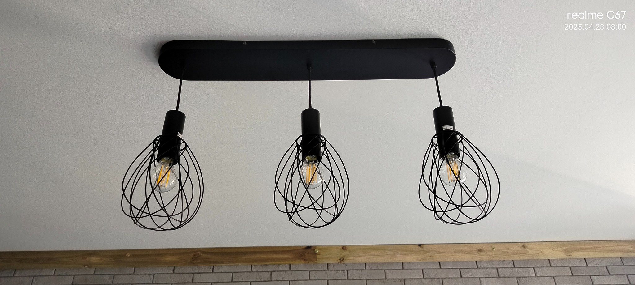 Czarna, designerska lampa sufitowa z trzema żarówkami w metalowych kloszach o nieregularnym kształcie, zamontowana na białym suficie. U dołu widoczny fragment ceglanej ściany.