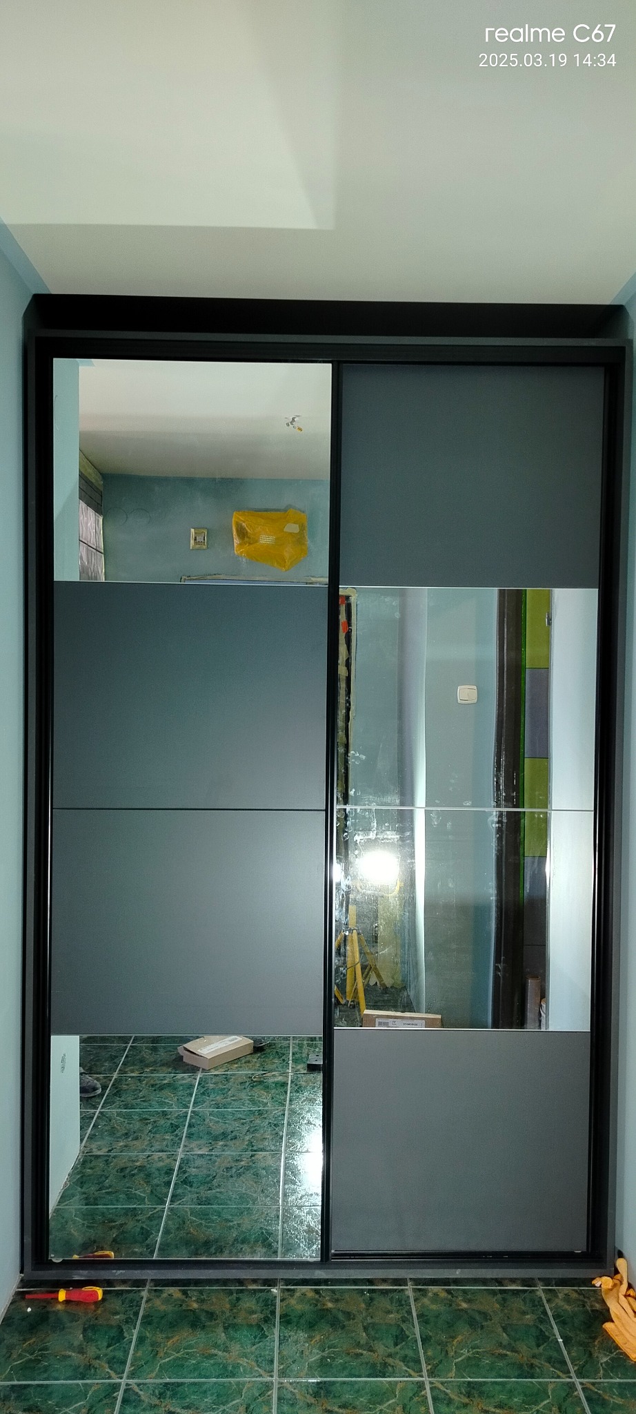 Szara szafa przesuwna z lustrami w mieszkaniu w trakcie remontu, widoczne narzędzia i zabezpieczone ściany. Ujęcie z bliska, wyraźny wzór płytek na podłodze.