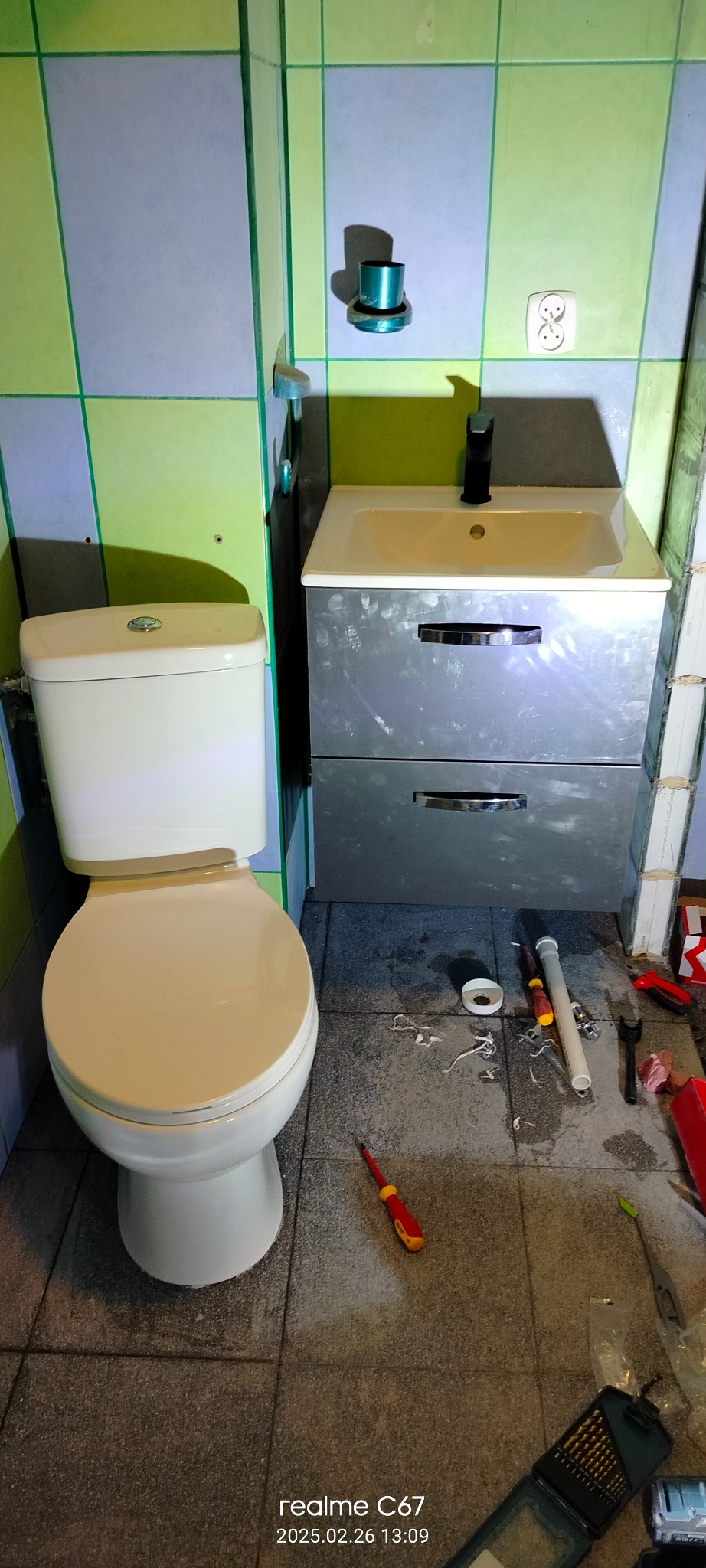 Montaż toalety i szafki z umywalką w łazience z zielono-niebieskimi płytkami. Na podłodze widoczne narzędzia instalacyjne i resztki materiałów.