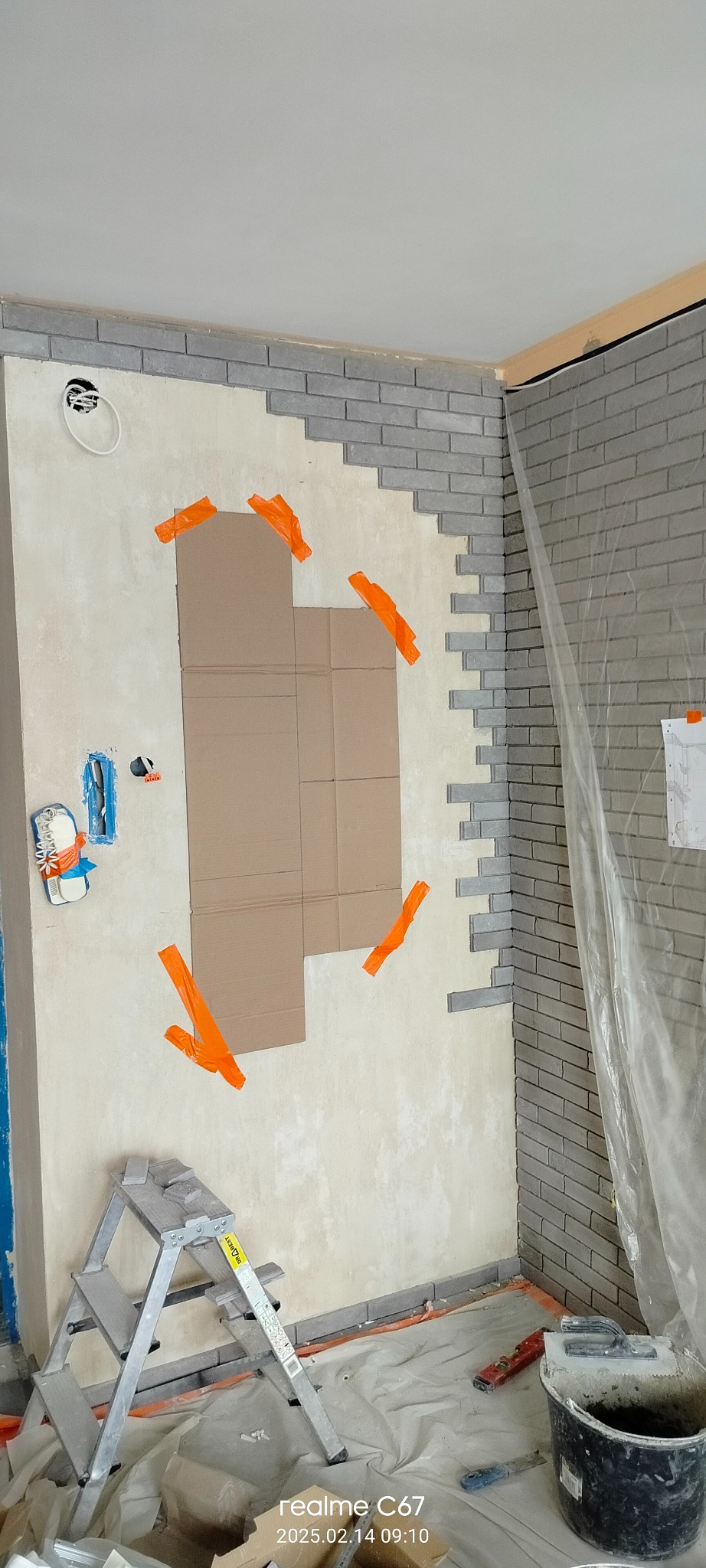 Wnętrze w trakcie prac: częściowo położona szara cegła dekoracyjna na ścianie, zabezpieczone kartonem miejsca pod gniazdka, drabina i narzędzia na podłodze.