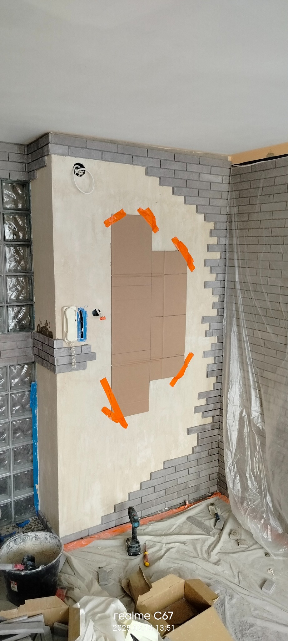 Montaż szarej cegły dekoracyjnej na ścianie, częściowo zakończony, z widocznymi narzędziami i zabezpieczeniami. Fragment instalacji elektrycznej i osłonięte szkło.