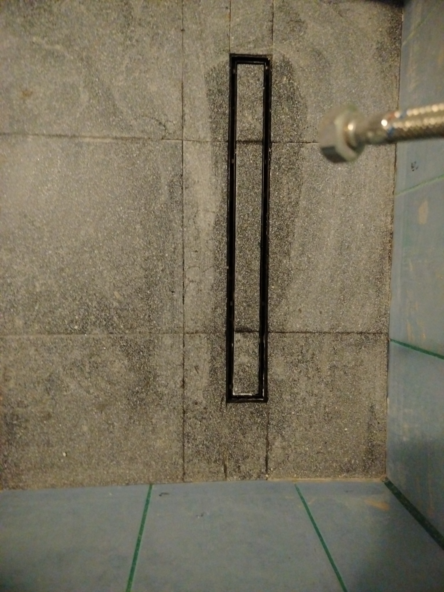 Szary brodzik z odpływem liniowym w trakcie montażu. Widoczny fragment wężyka doprowadzającego wodę. Płytki imitujące kamień.