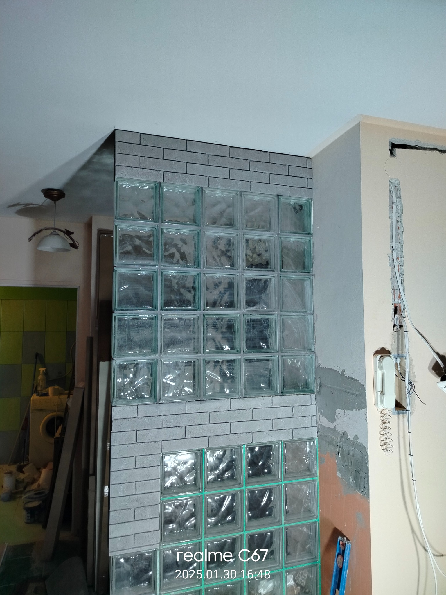 Ściana z luksferów w trakcie montażu, obłożona szarą cegłą. Widoczne niedociągnięcia i przewody elektryczne. W tle fragment łazienki z pralką.