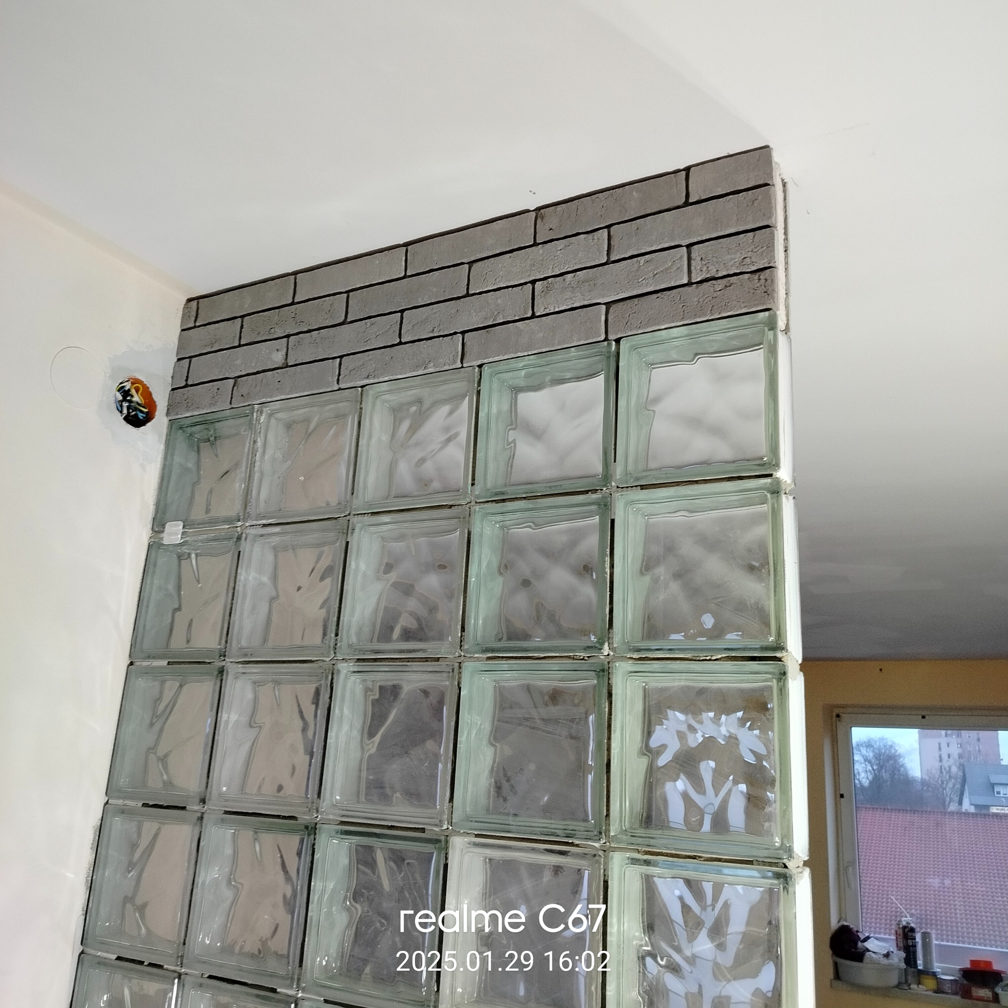 Ścianka z luksferów i cegieł w szarej tonacji, widok z boku, z widocznym fragmentem instalacji elektrycznej w ścianie. Widoczne okno w tle.