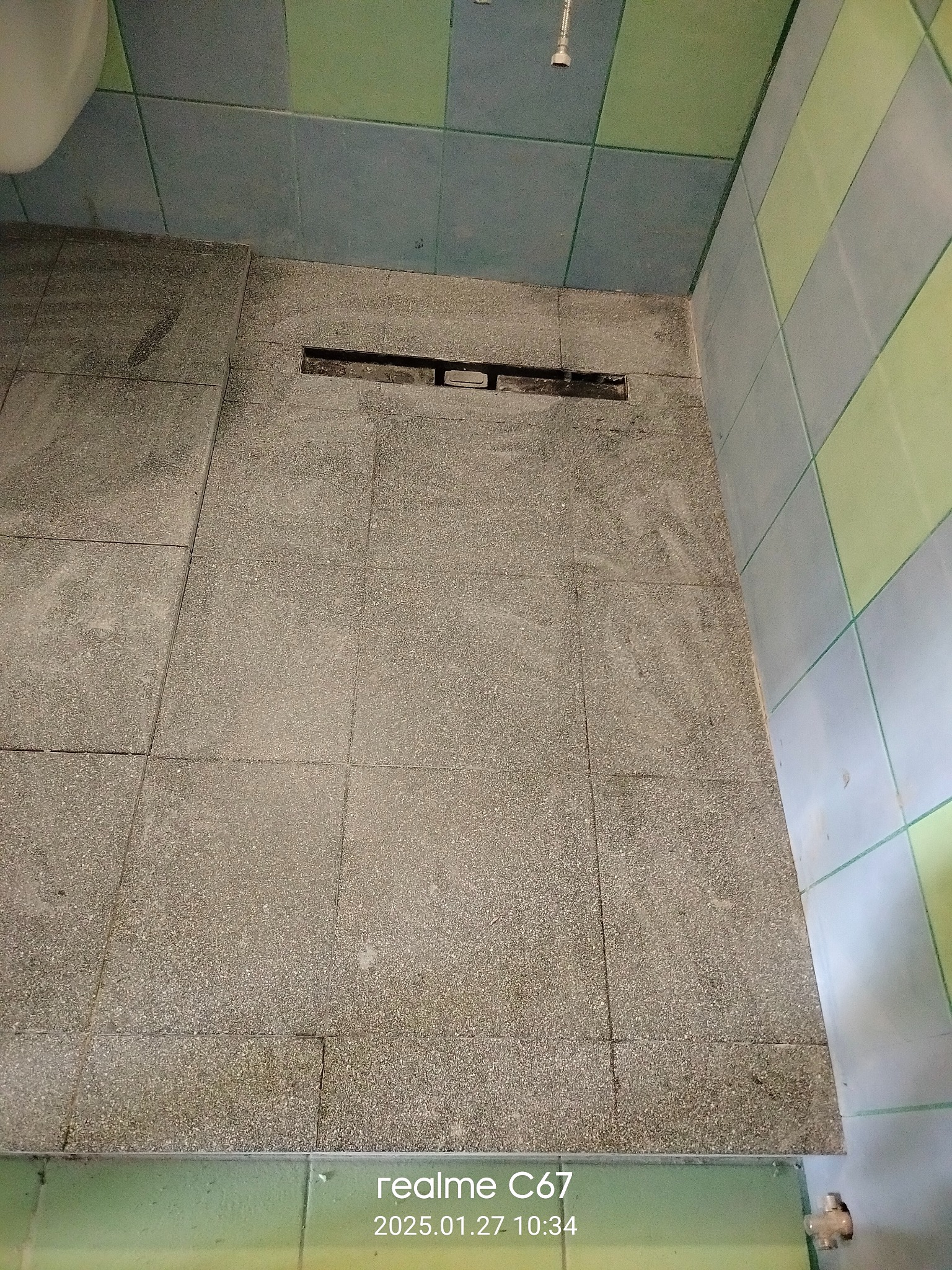 Ukończona podłoga w łazience z szarych płytek i odpływem liniowym, widok z góry. Fragment toalety i kolorowe płytki na ścianie.