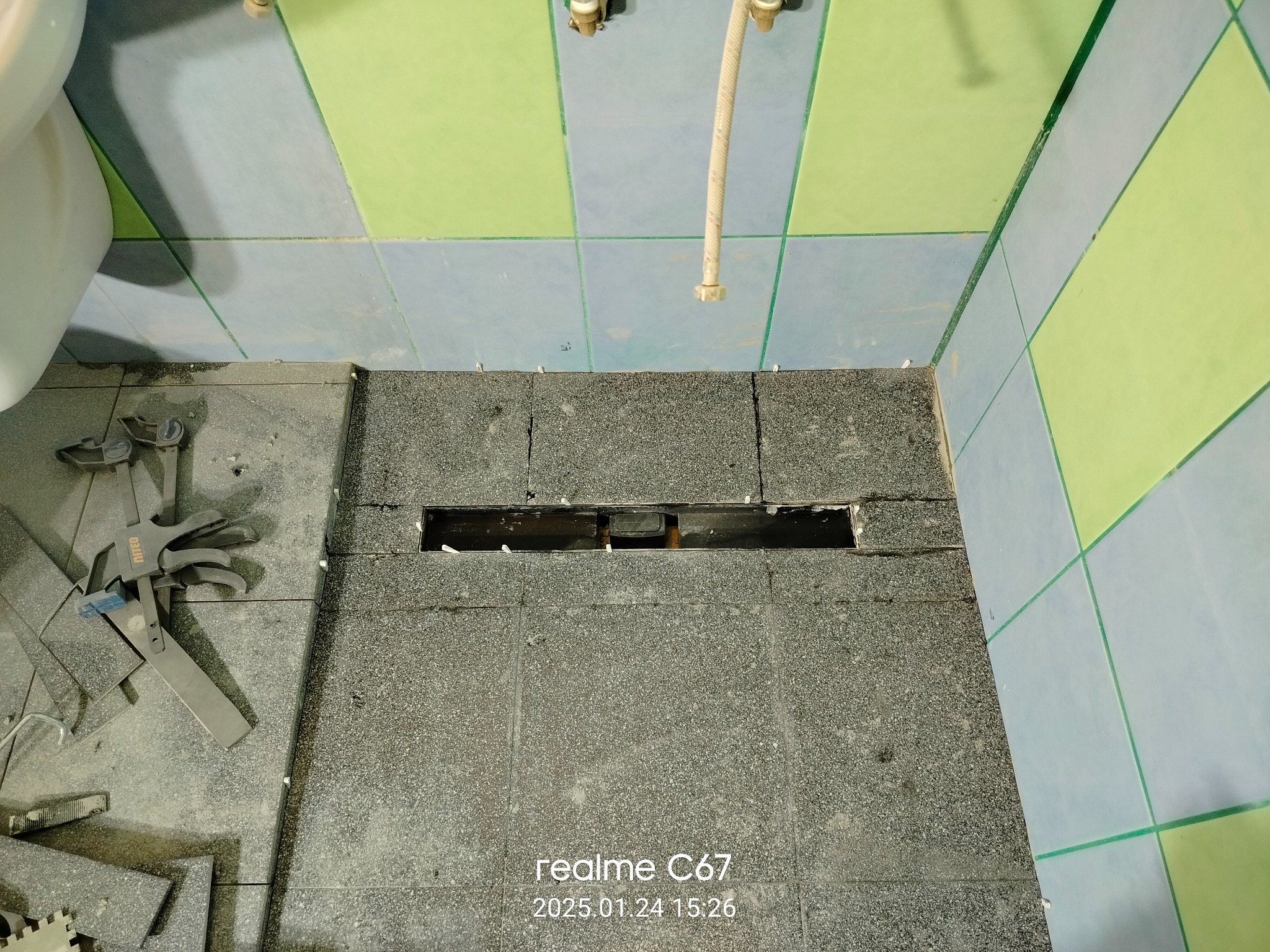 Szare płytki podłogowe w łazience w trakcie układania, widoczne narzędzia i fragment instalacji. Ściany z zielono-niebieskimi kaflami.