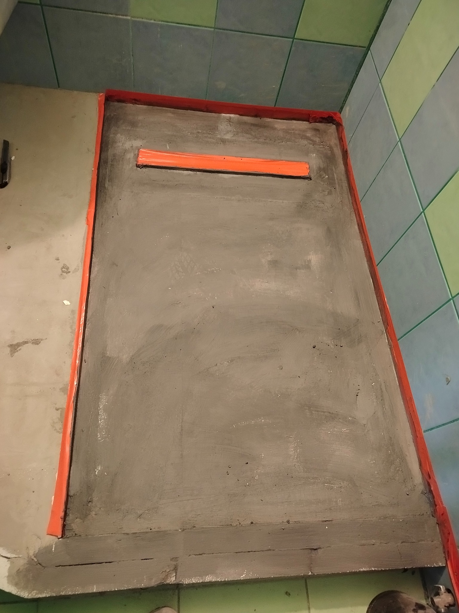 Szary brodzik z odpływem liniowym w pomarańczowej ramce, widoczny fragment łazienki z zielono-niebieskimi płytkami. Etap prac remontowych.