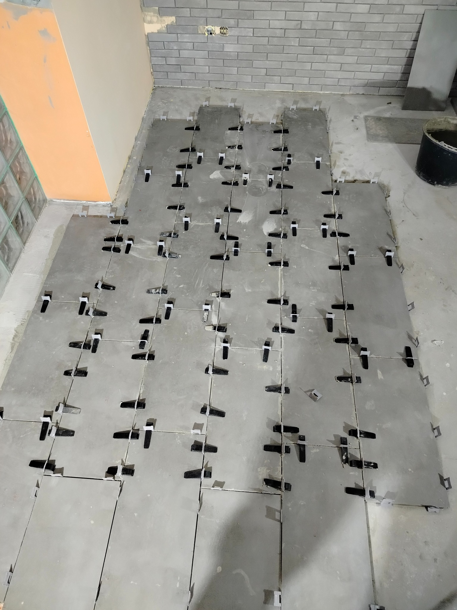 Szare płytki podłogowe w trakcie układania, z czarnymi klipsami poziomującymi, widoczne fragmenty ścian z różnym wykończeniem.