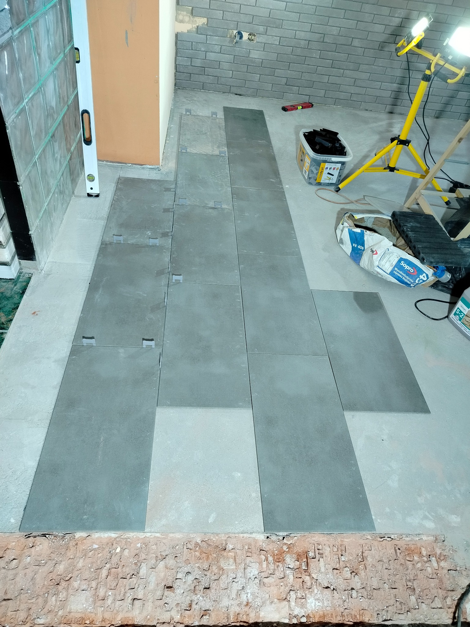 Układanie szarych płytek podłogowych z użyciem klipsów dystansowych w Płocku. Widoczny poziomica, lampa budowlana i zaprawa klejowa.