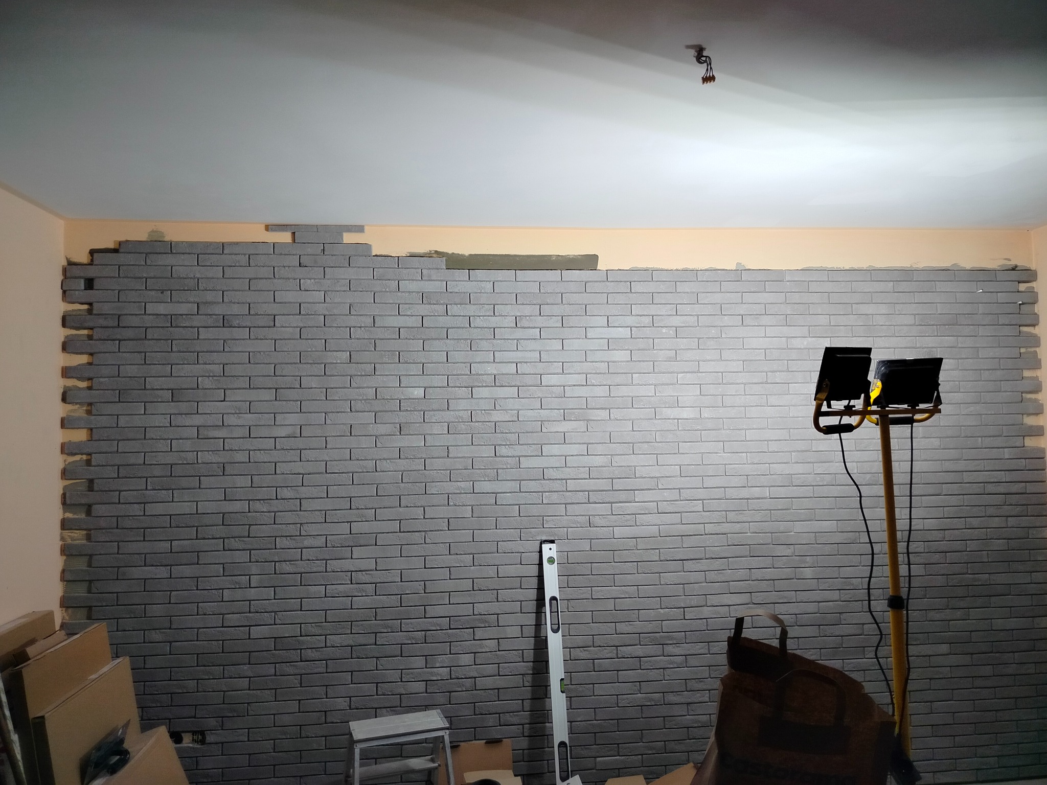 Montaż szarej okładziny imitującej cegłę na ścianie, widoczny poziomica, lampa oświetleniowa i kartony. Prace w toku.