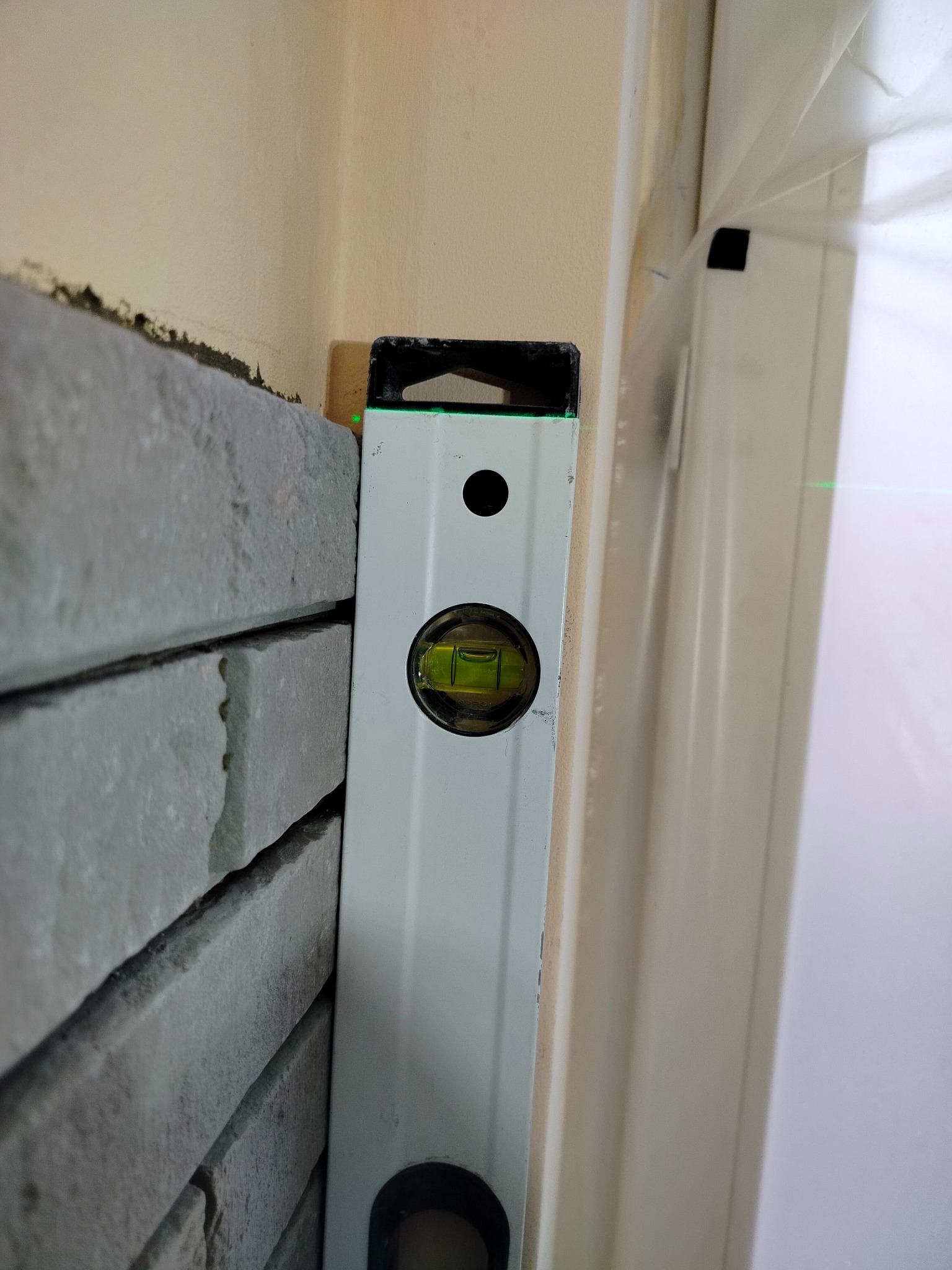 Poziomica przy szarej okładzinie ściennej w rogu pokoju, obok okna z folią ochronną. Detal z prac wykończeniowych.