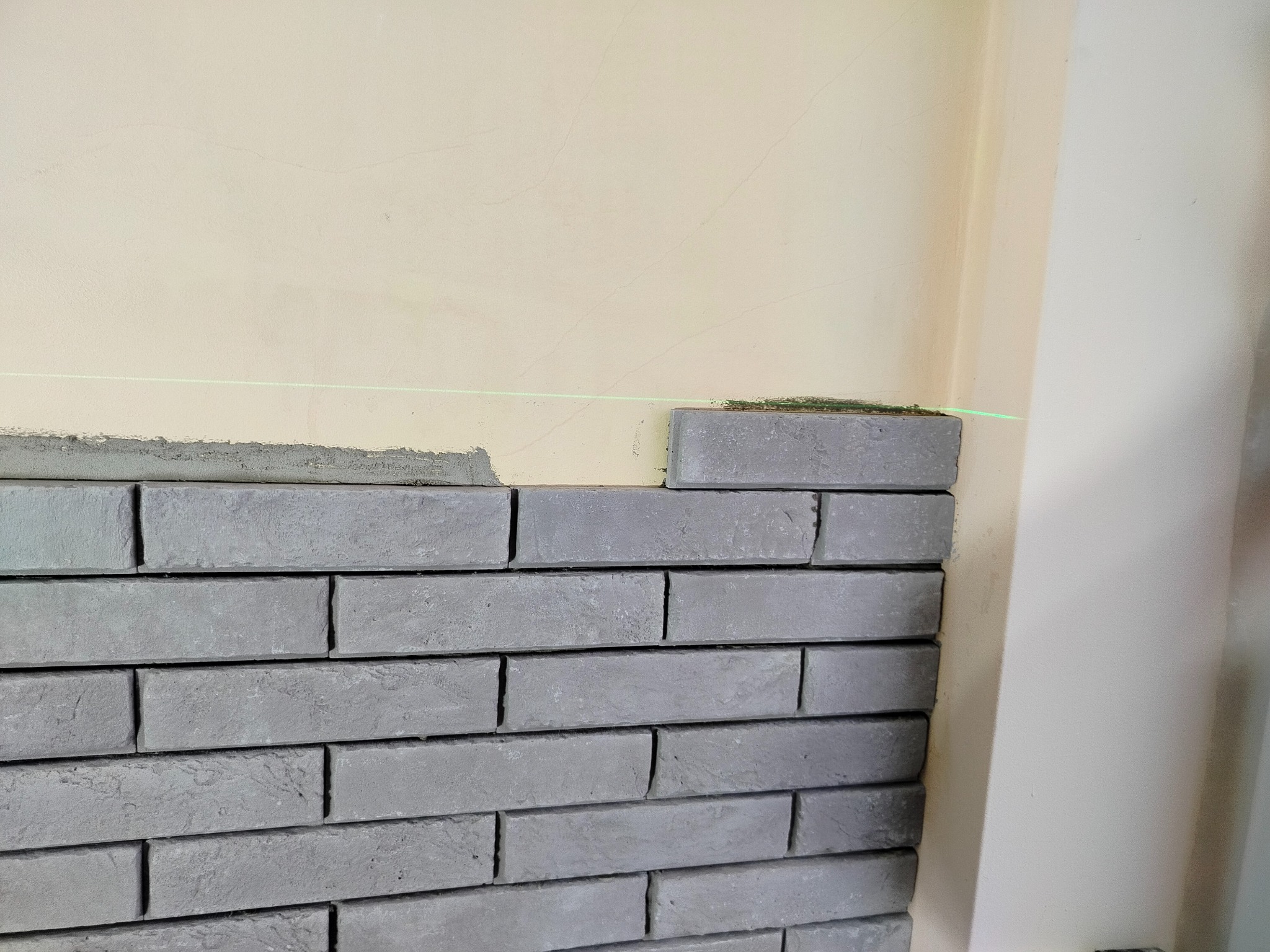 Fragment ściany z szarej cegły dekoracyjnej, układanej równo, z użyciem poziomicy laserowej, na tle kremowej ściany. Widoczny etap prac wykończeniowych.