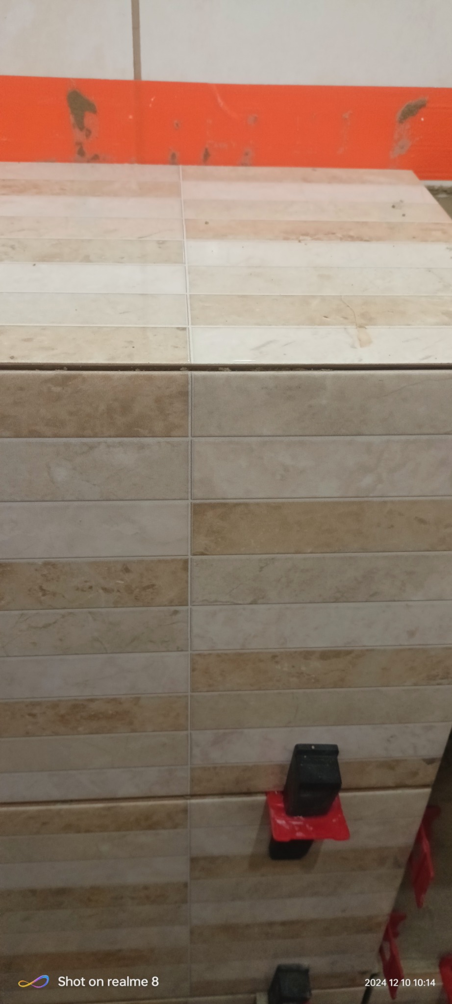 Ściana wyłożona beżowo-brązowymi, prostokątnymi płytkami ceramicznymi, z użyciem klipsów poziomujących. Widoczne fugi i fragment pomarańczowej taśmy malarskiej u góry.