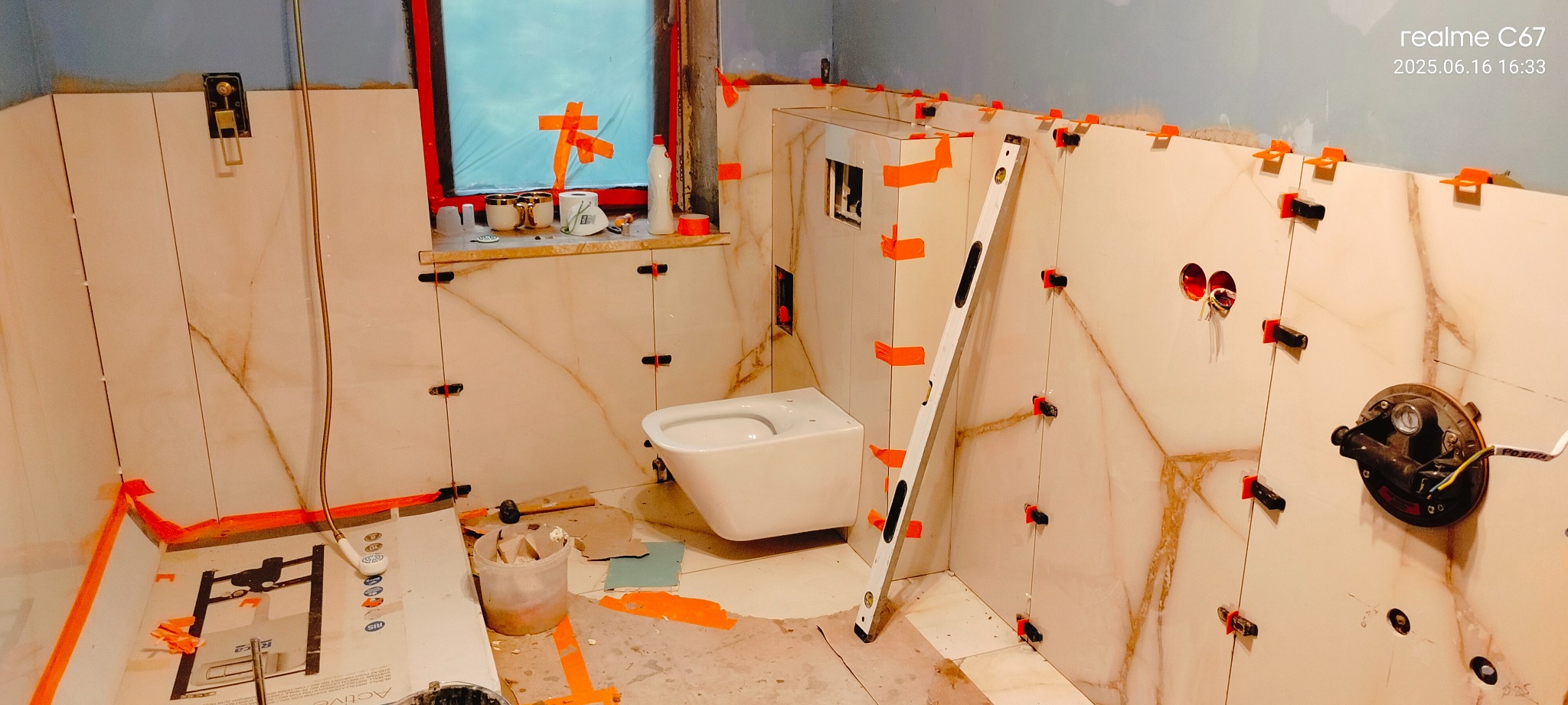 Remont łazienki w trakcie: białe płytki imitujące marmur, poziomica, toaleta podwieszana, okno zabezpieczone folią. Widoczne narzędzia i materiały budowlane.