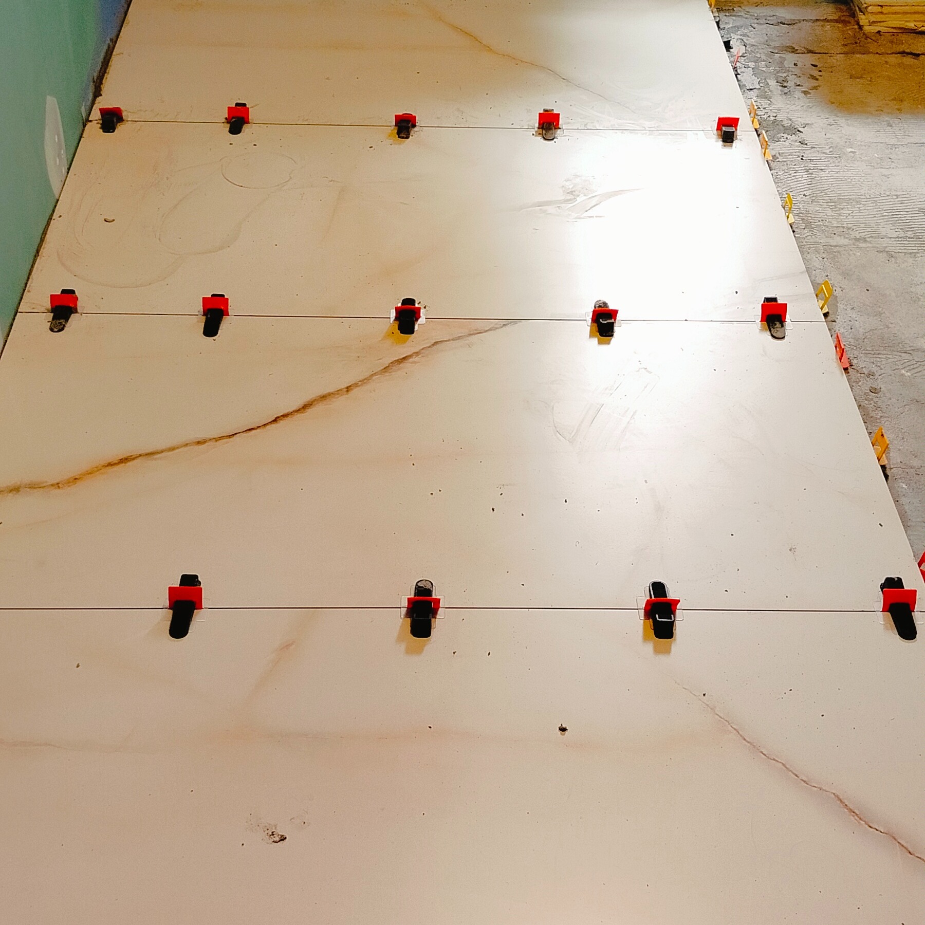 Układanie płytek podłogowych z użyciem systemu poziomowania. Widoczne płytki imitujące marmur z delikatnym wzorem i czerwone klipsy dystansowe.