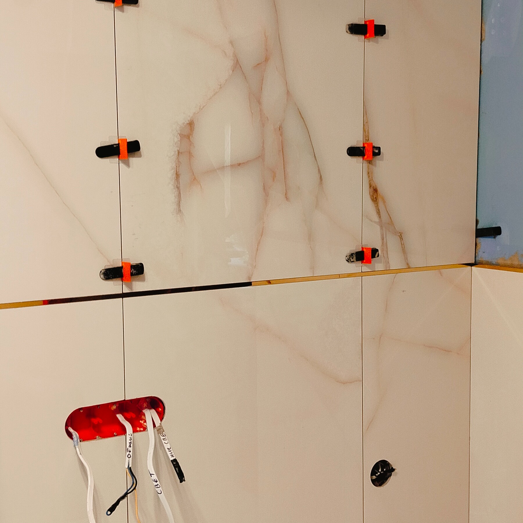 Montaż płytek imitujących marmur z użyciem klipsów poziomujących. Widoczne przewody elektryczne w puszce. Detal wykończenia łazienki w trakcie prac.