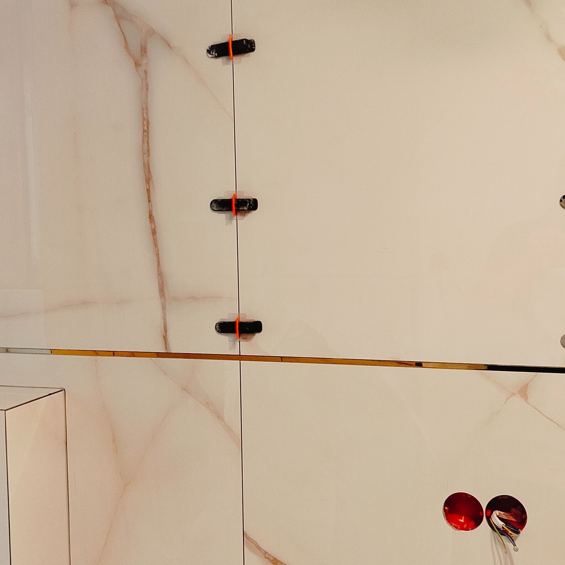 Ściana wyłożona białymi płytkami imitującymi marmur z pomarańczowymi żyłkami, z widocznymi klipsami poziomującymi i otworami na gniazdka elektryczne.