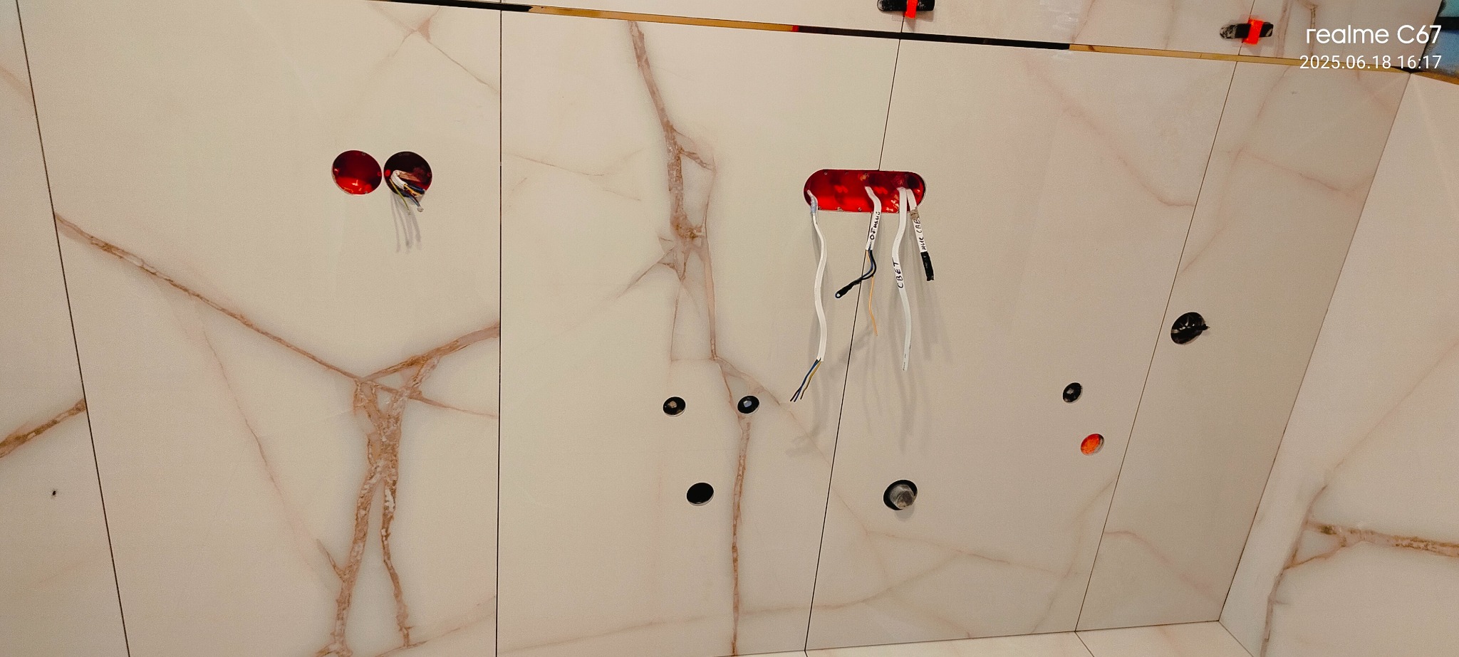 Wykończona ściana z marmurowych płytek z widocznymi otworami pod gniazdka i wyprowadzonymi przewodami elektrycznymi. Widoczne czerwone puszki instalacyjne.