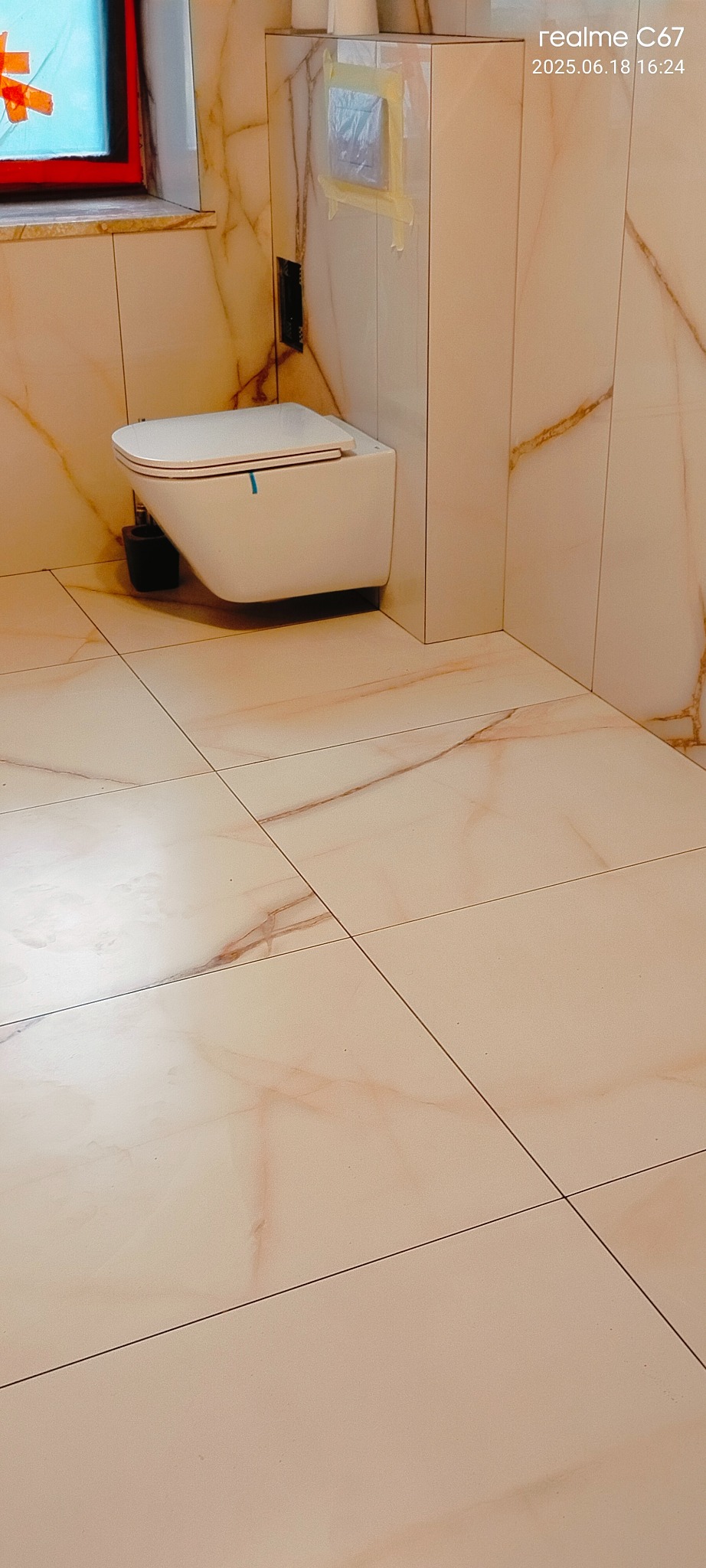 Wykończona łazienka z podwieszaną toaletą na tle marmurowych płytek. Widoczny fragment okna z pomarańczową ramą i zaklejony taśmą przycisk spłuczki.