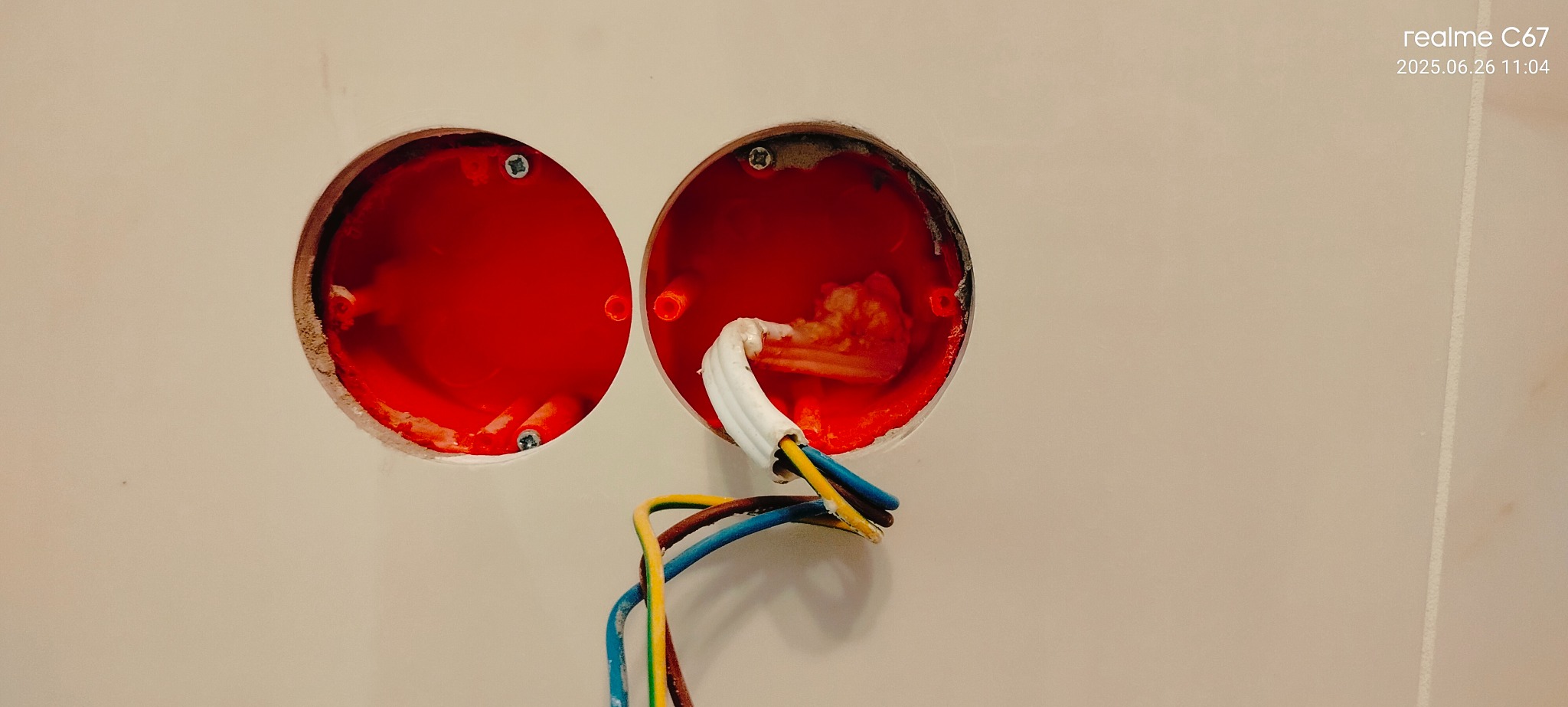 Dwa czerwone, okrągłe otwory w ścianie z wystającymi przewodami elektrycznymi. Widoczne ślady po szpachlowaniu wokół otworów. Ujęcie z bliska, oświetlenie naturalne.