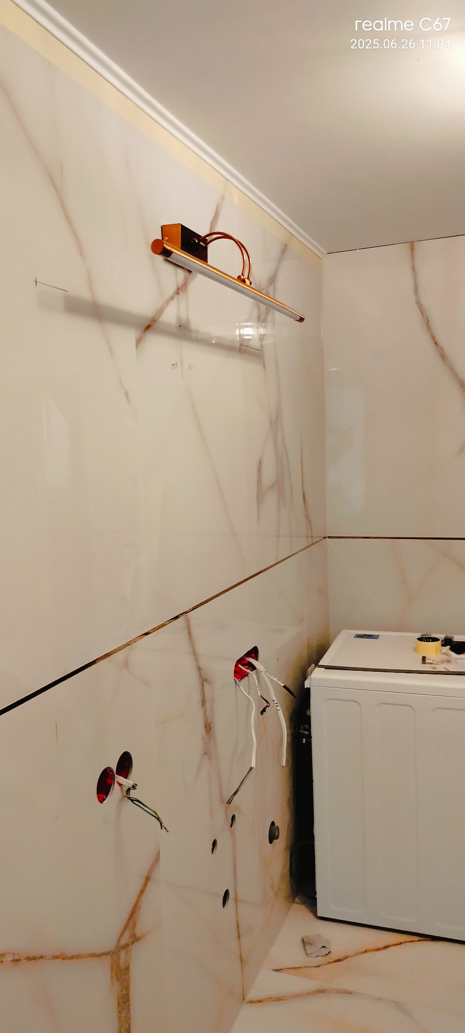 Łazienka w trakcie wykańczania: marmurowe płytki, zamontowana lampa nad lustrem, widoczne otwory na gniazdka elektryczne i pralka. Etap prac wykończeniowych.