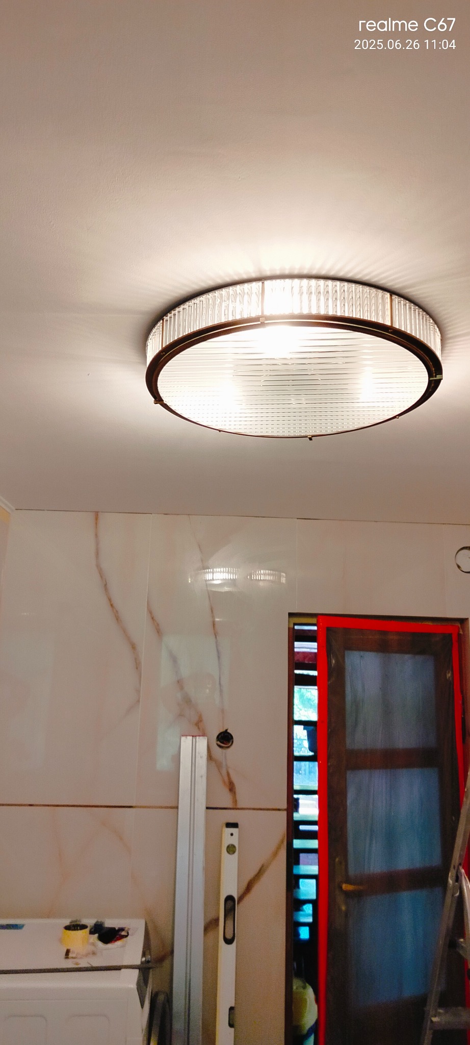 Zamontowana okrągła lampa sufitowa z kryształkami w łazience z marmurowymi płytkami. Widoczne narzędzia i poziomica, sugerujące prace wykończeniowe.