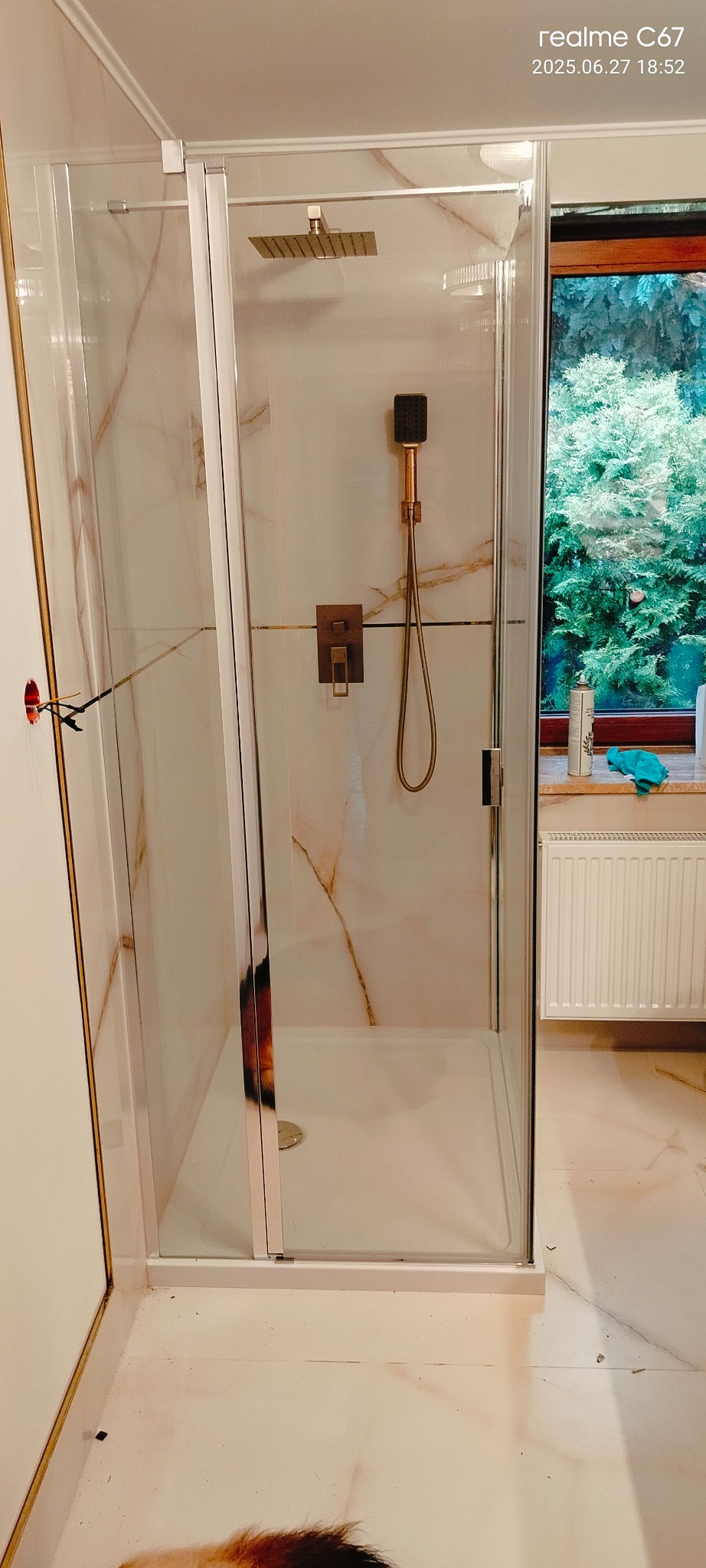 Nowoczesna kabina prysznicowa z deszczownicą i baterią w odcieniach brązu, wykończona marmurem. Widok na okno z zielenią w tle. Elementy instalacji elektrycznej widoczne.