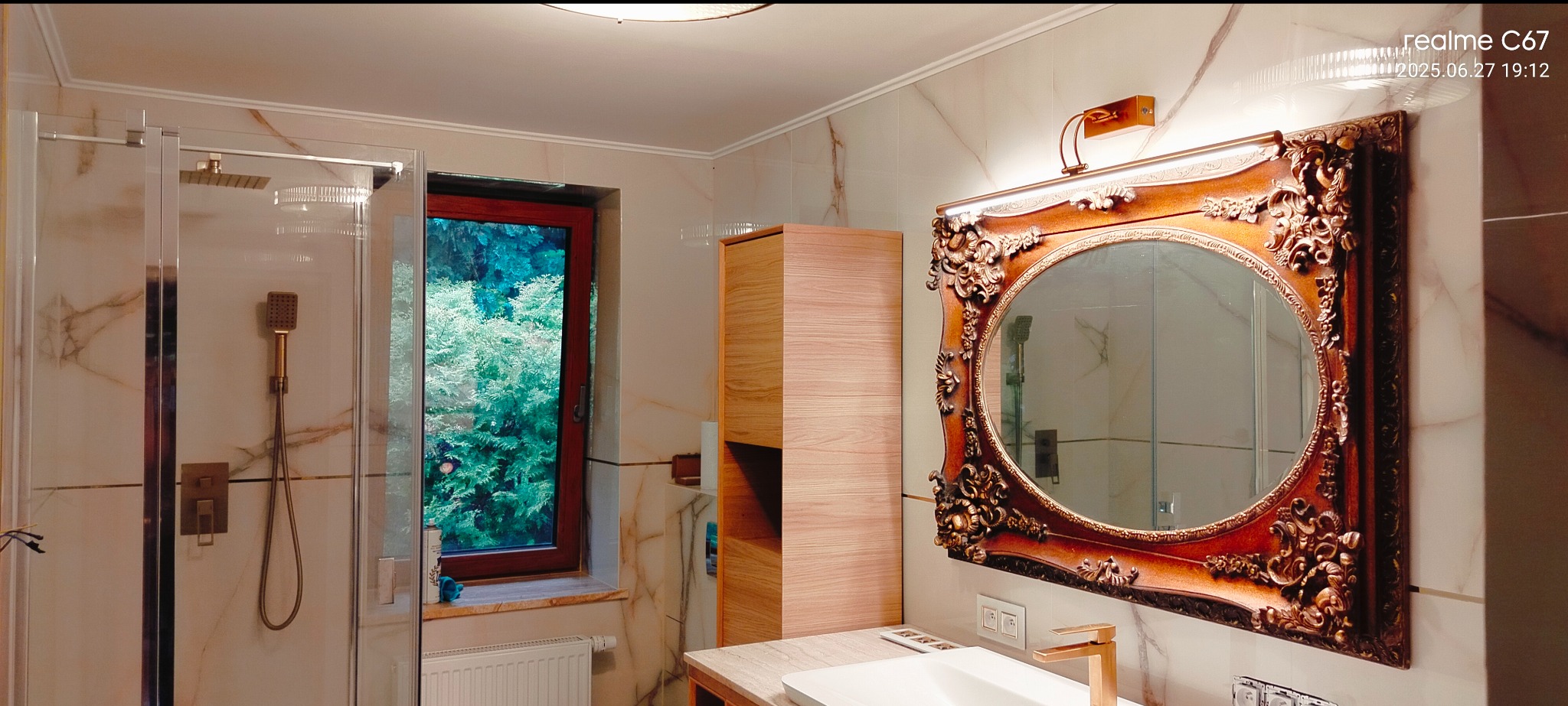 Elegancka łazienka z kabiną prysznicową, oknem z widokiem na zieleń i ozdobnym lustrem w złotej ramie. Marmurowe płytki na ścianach, złota armatura.