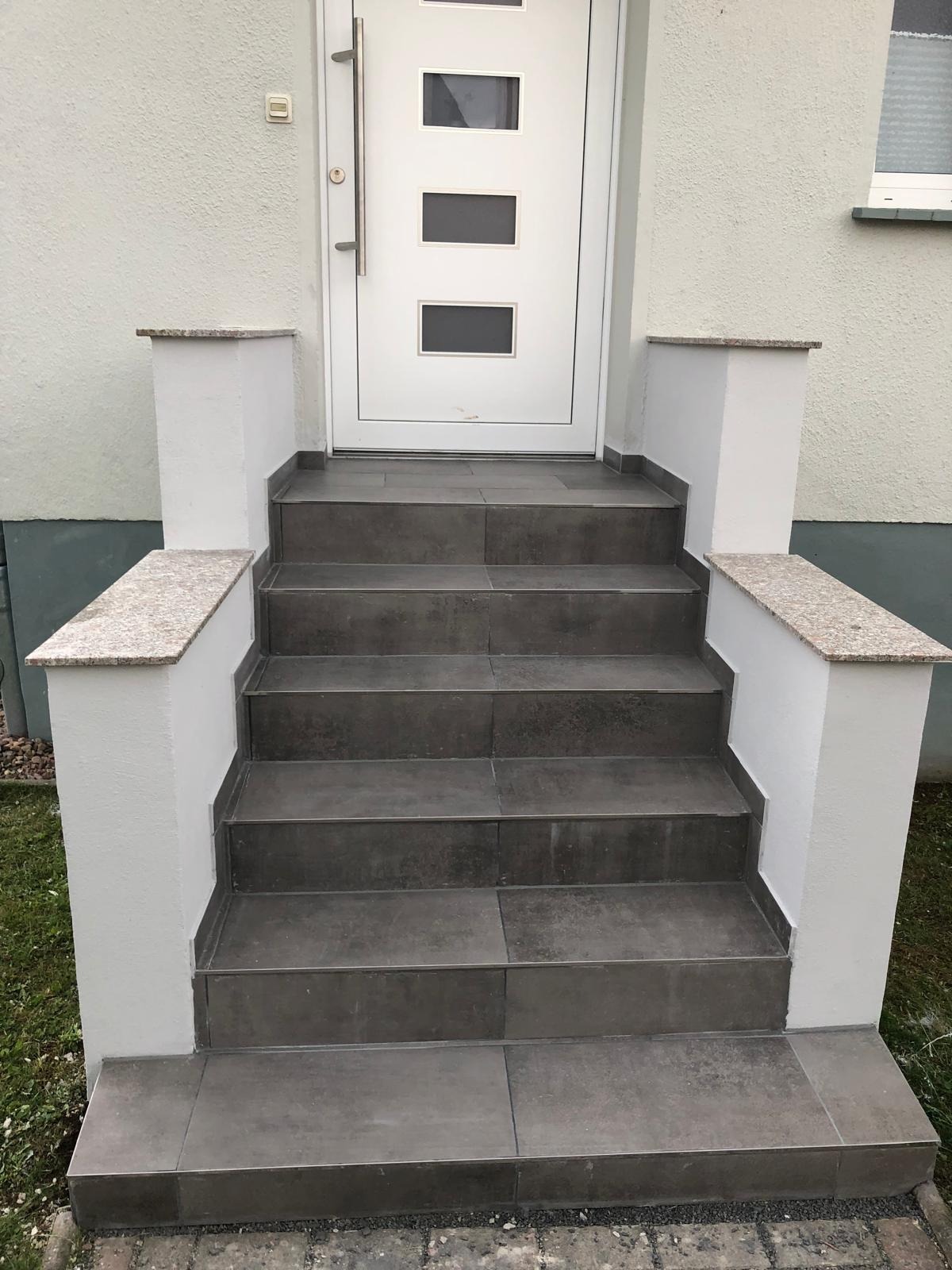 Zewnętrzne schody wejściowe wyłożone szarymi płytkami, z białymi słupkami z granitowymi nakrywami, prowadzące do białych drzwi z trzema prostokątnymi okienkami.