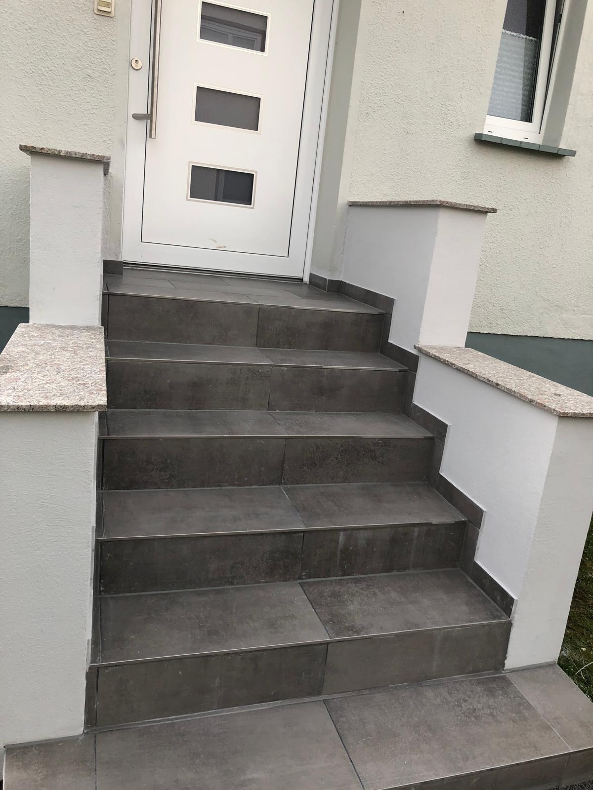 Zewnętrzne schody wejściowe wyłożone ciemnoszarą płytką, z białymi ściankami bocznymi zwieńczonymi płytami z jasnego granitu, prowadzące do białych drzwi wejściowych z trzema prostokątnymi okienkami.