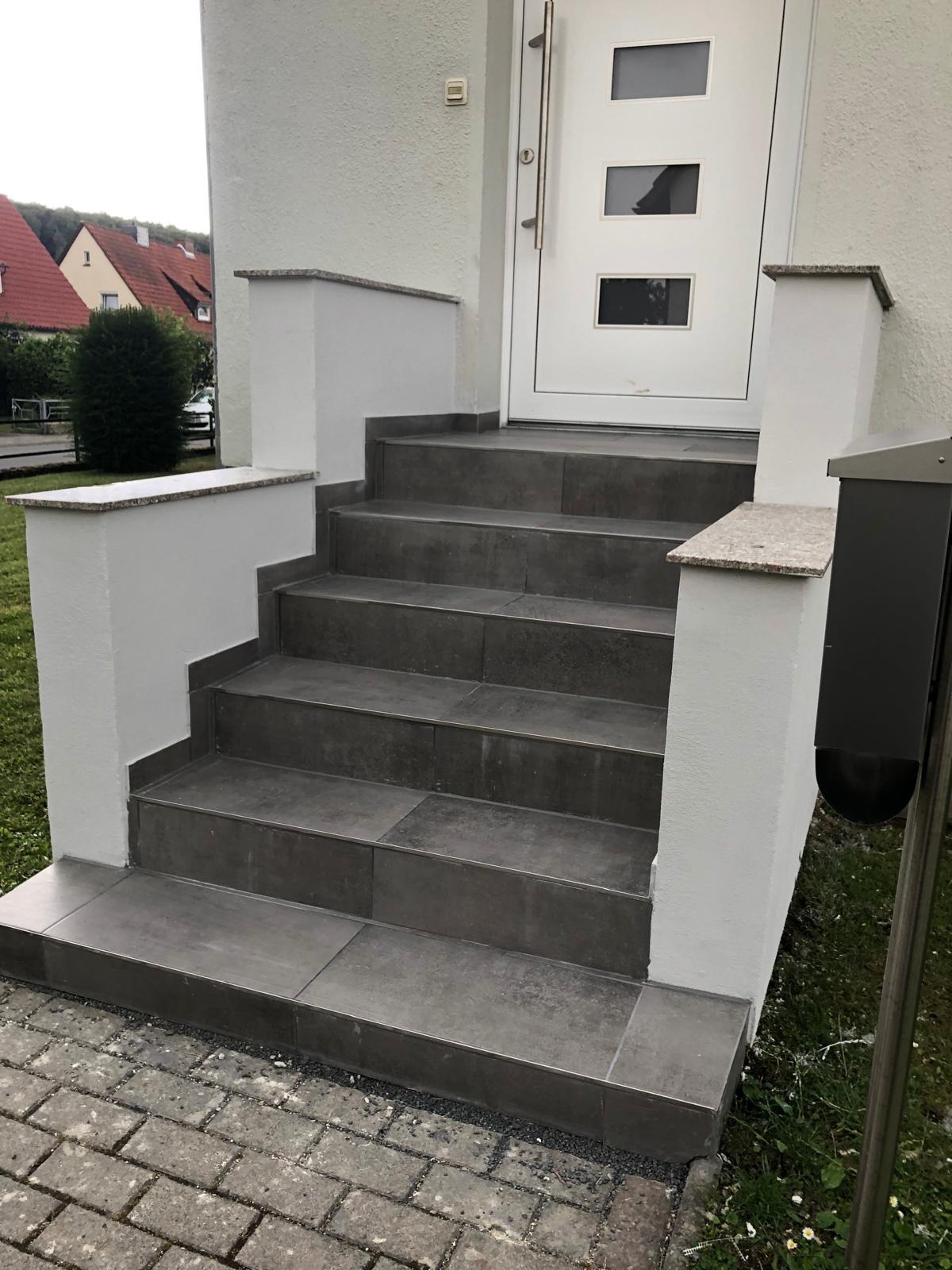 Zewnętrzne schody wejściowe wyłożone szarą płytką, z białymi ściankami bocznymi zwieńczonymi jasnym granitem, prowadzące do białych drzwi z trzema szybami.