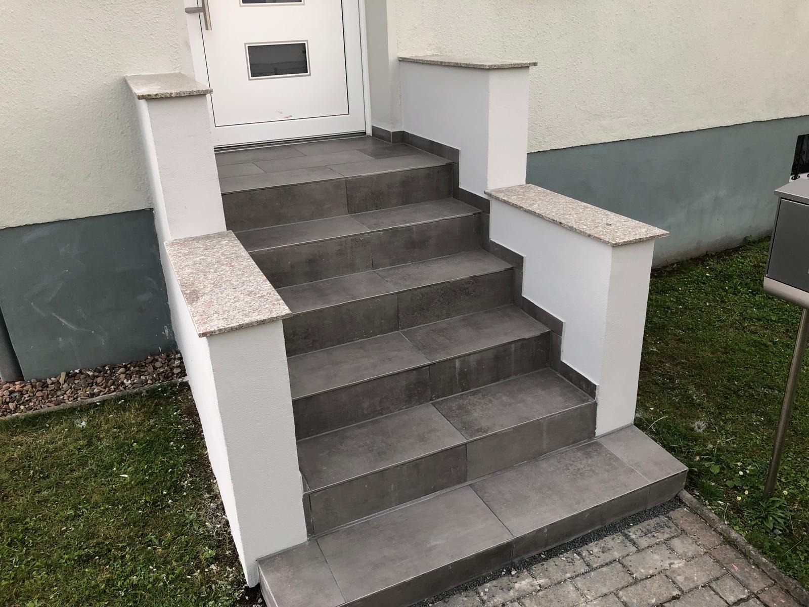 Zewnętrzne schody wejściowe wyłożone szarymi płytkami, z białymi ściankami bocznymi zwieńczonymi jasnym granitem, prowadzące do białych drzwi.