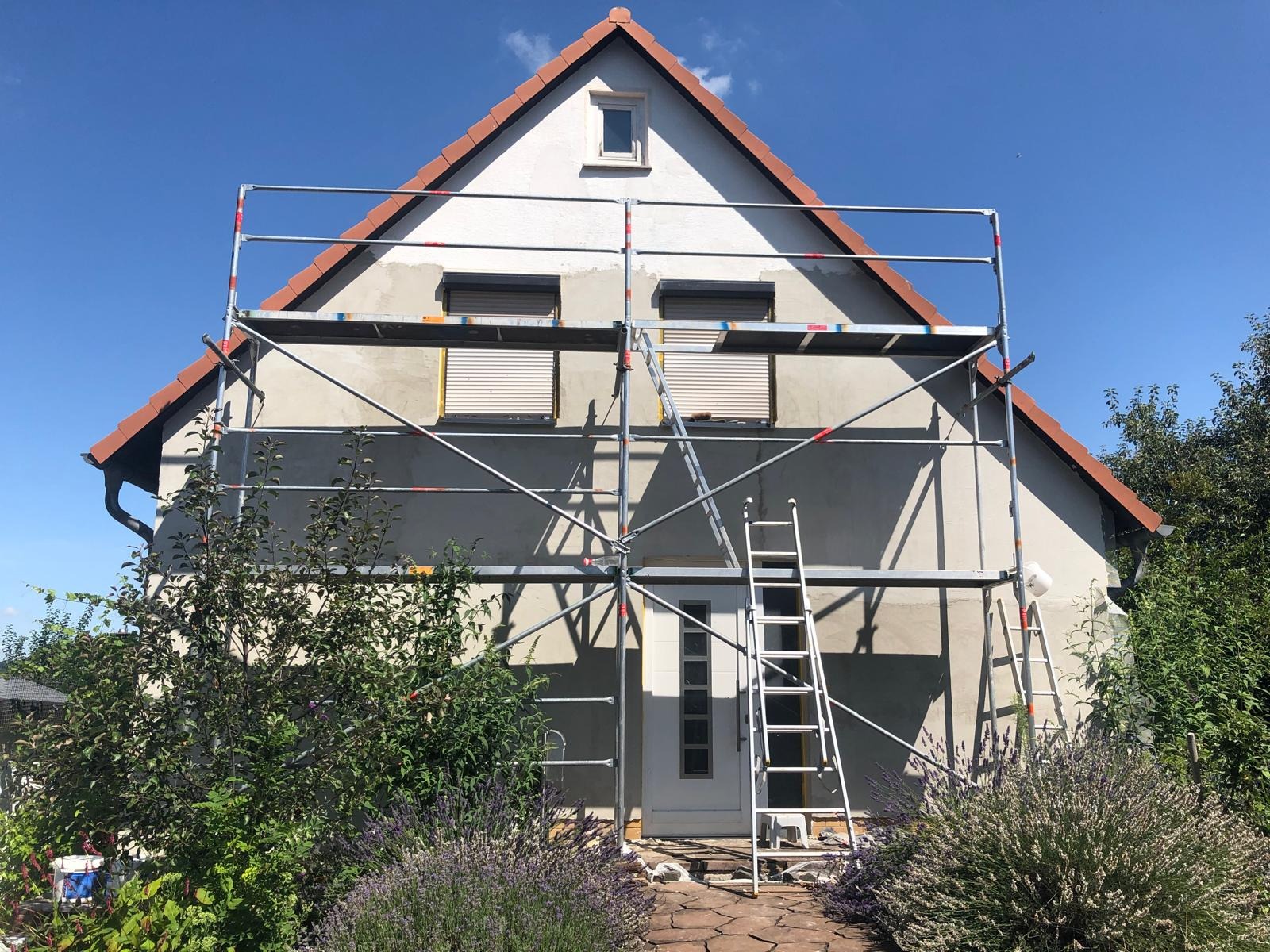 Dom jednorodzinny w trakcie przygotowania do malowania elewacji, z widocznym rusztowaniem i zabezpieczonymi oknami, na tle błękitnego nieba.