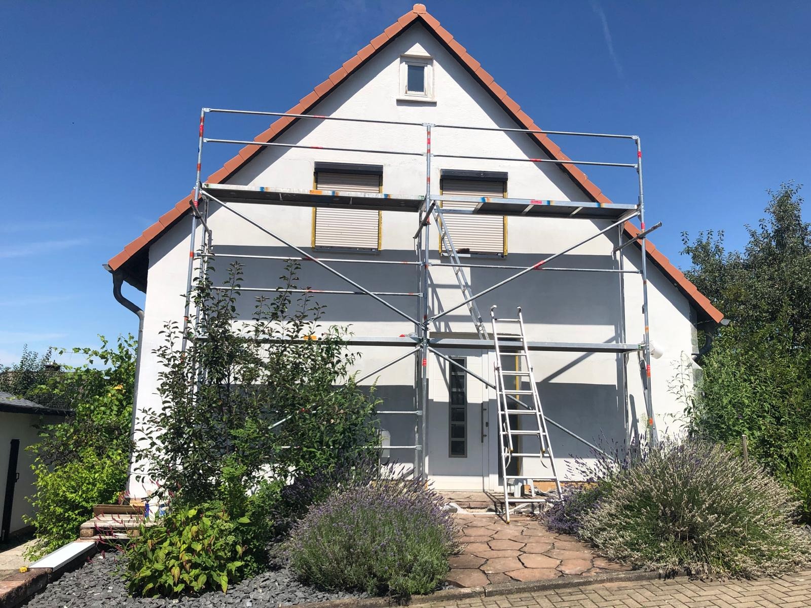 Dom jednorodzinny w trakcie malowania elewacji, z widocznym rusztowaniem i częściowo pomalowaną ścianą na szaro, jasne rolety w oknach, drabina oparta o ścianę.