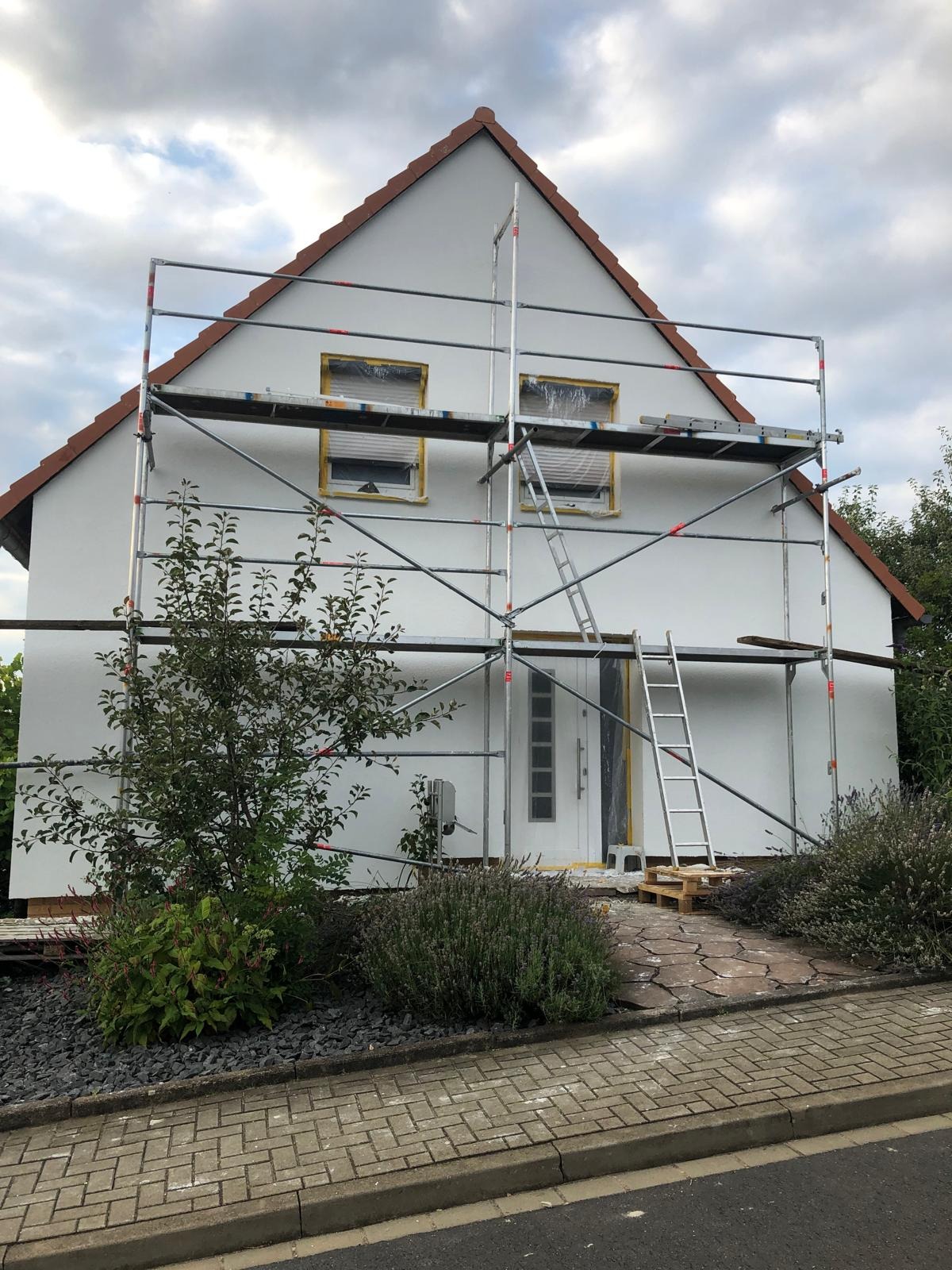 Dom jednorodzinny w trakcie malowania elewacji, zabezpieczone okna taśmą malarską, rusztowanie rozstawione, drabina oparta o elewację.