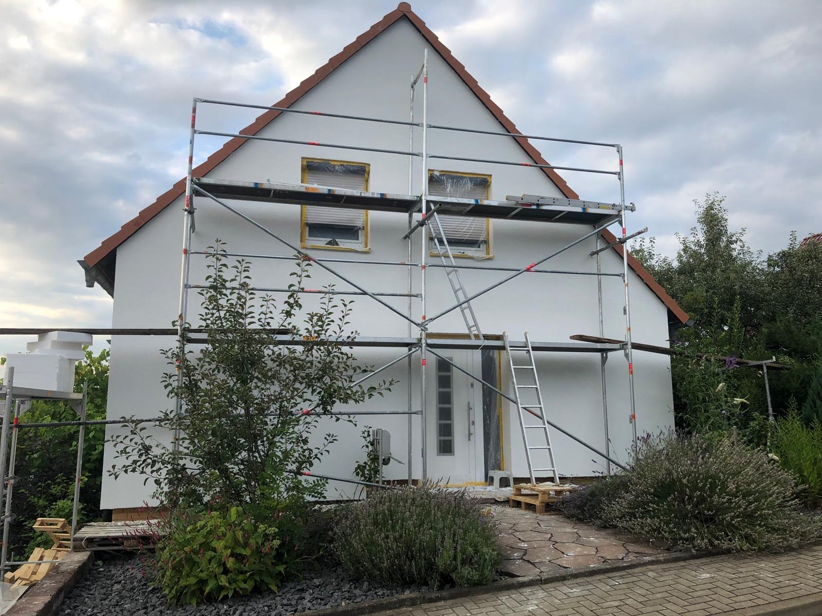 Dom jednorodzinny w trakcie odświeżania elewacji, zabezpieczone okna folią malarską, rozstawione rusztowanie, widoczne ślady farby na elewacji, podłoże z kamieni i kostki brukowej.