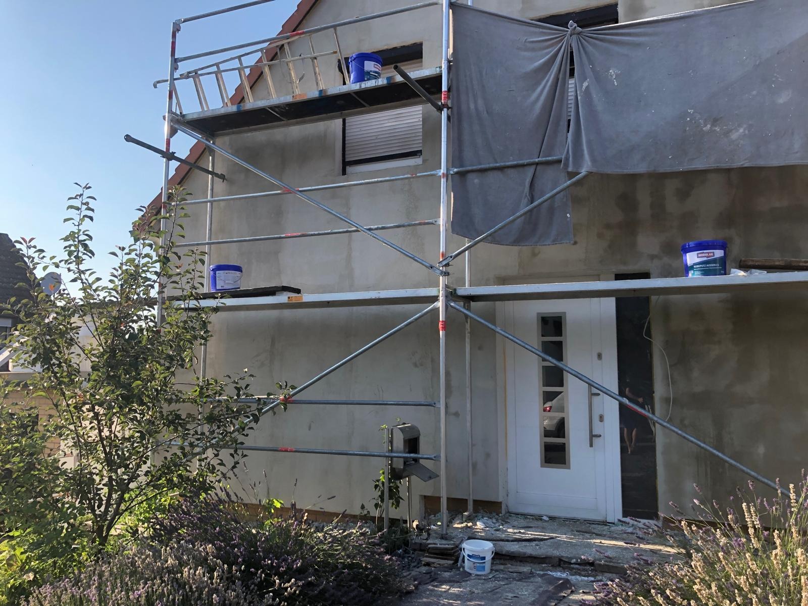 Dom w trakcie przygotowania do malowania elewacji, widoczne rusztowanie, wiaderka z farbą i zabezpieczona okładzina. W tle drzewo i krzewy.